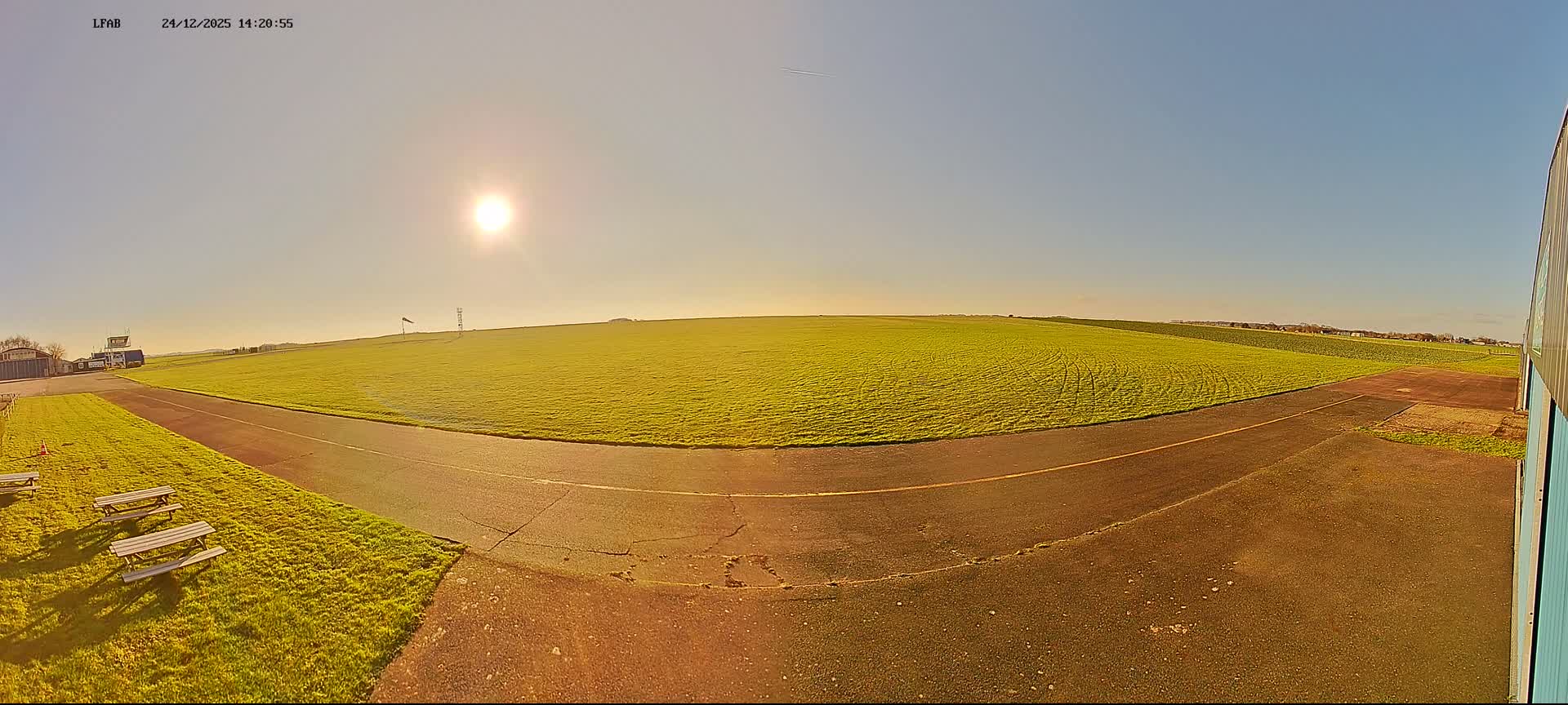 An overcast sky hangs over a panoramic view of a small airfield, featuring a vast green grass field dotted with white objects, a winding paved path, several distant buildings including a control tower, and three picnic tables in the foreground.