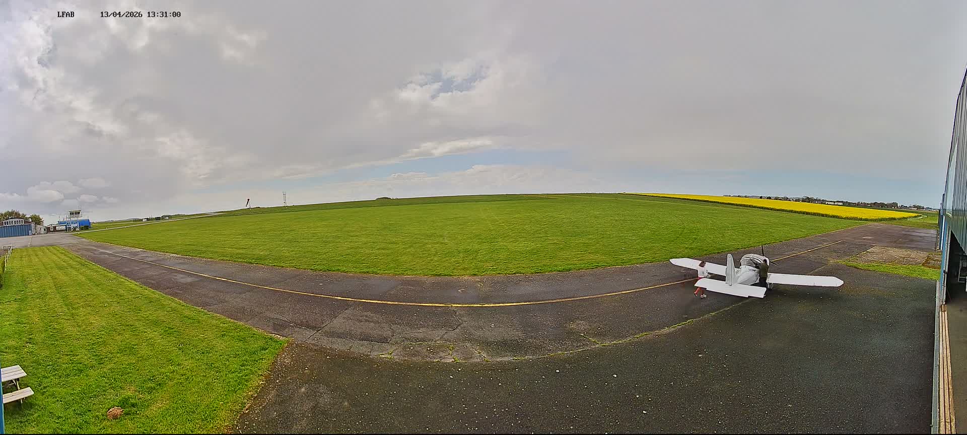 An overcast sky hangs over a panoramic view of a small airfield, featuring a vast green grass field dotted with white objects, a winding paved path, several distant buildings including a control tower, and three picnic tables in the foreground.