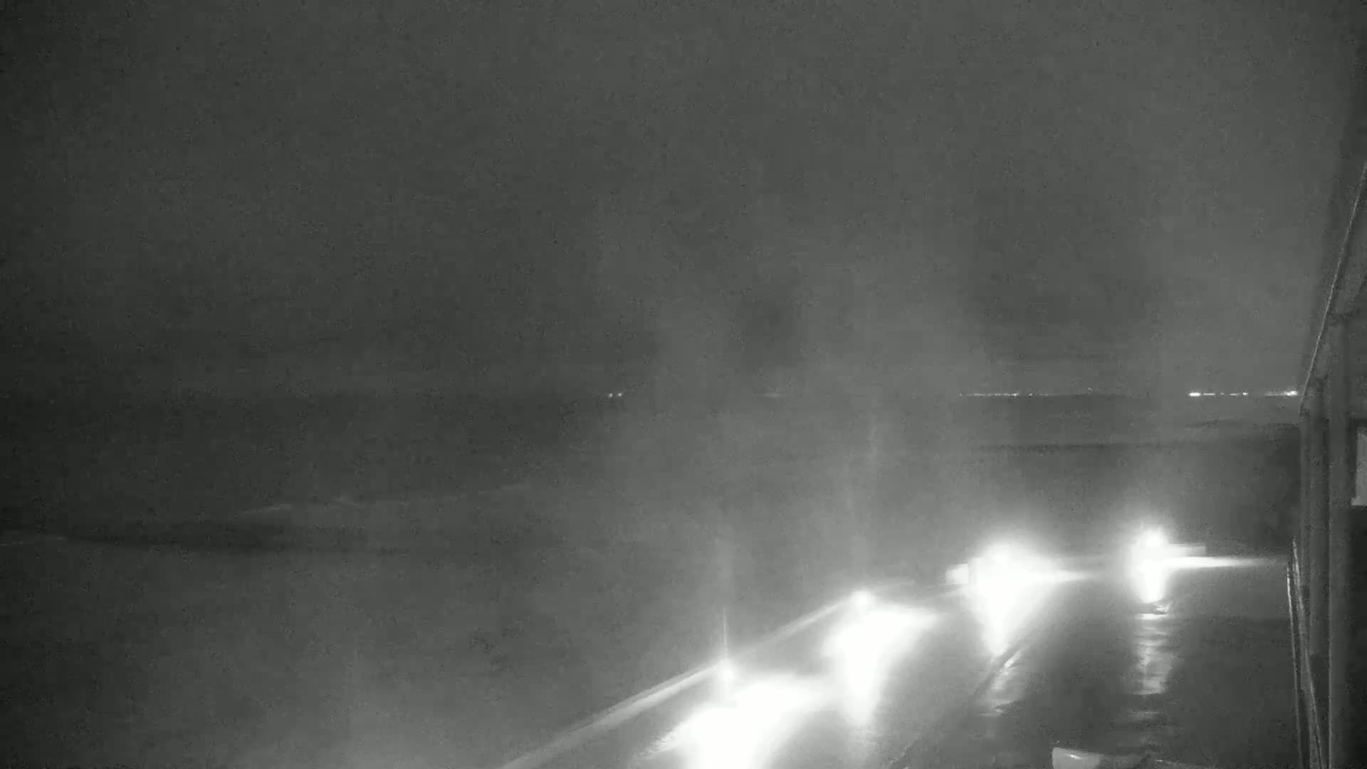 A nighttime, foggy scene shows a wet road with headlights visible in the distance, and a building partially visible on the right.