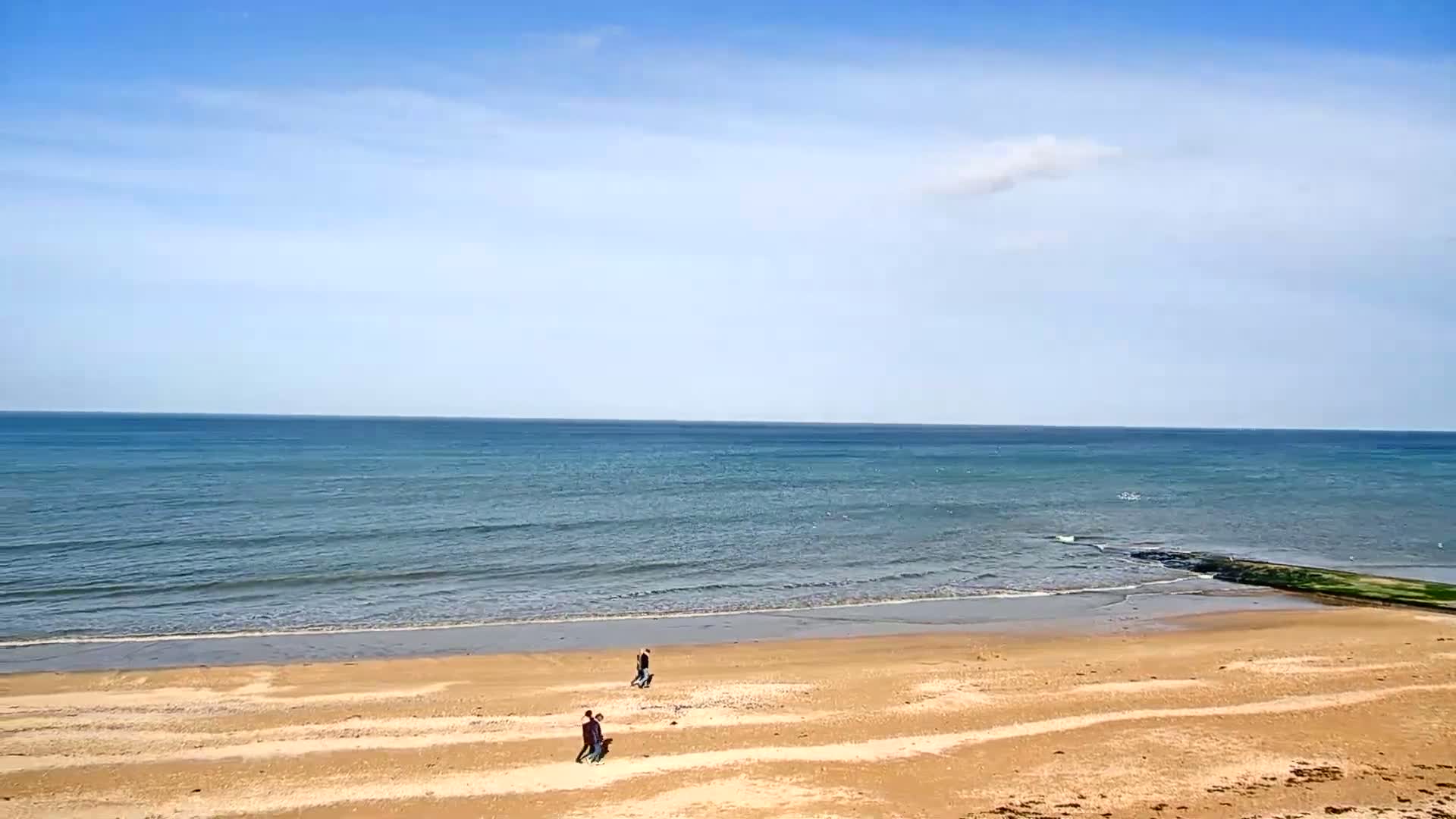 Under a mostly clear blue sky, two pairs of people stroll along a wide sandy beach that slopes gently down to a calm, blue-green sea.