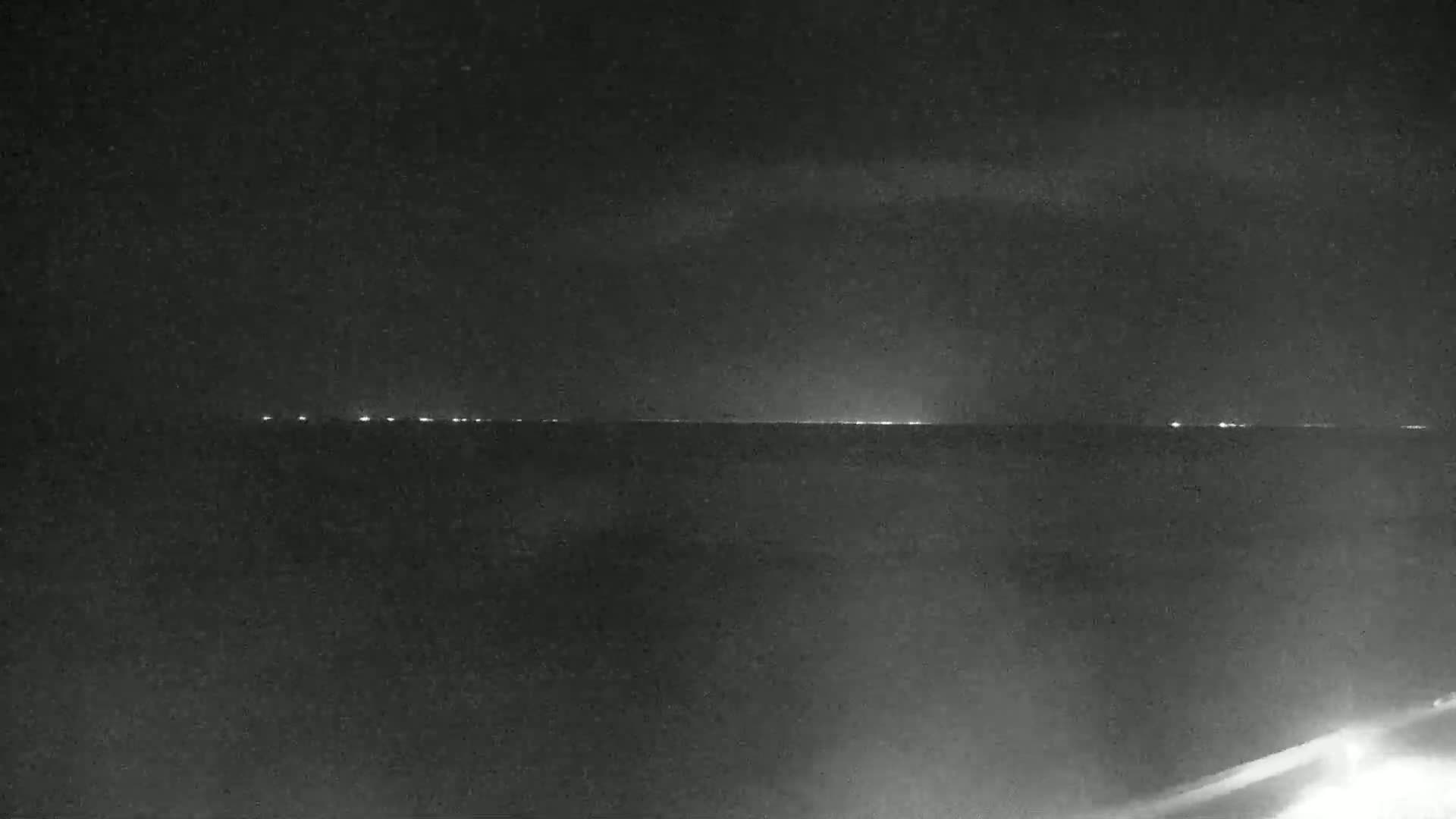 A nighttime view of a dark body of water with a faint line of lights on the horizon under a mostly clear sky.