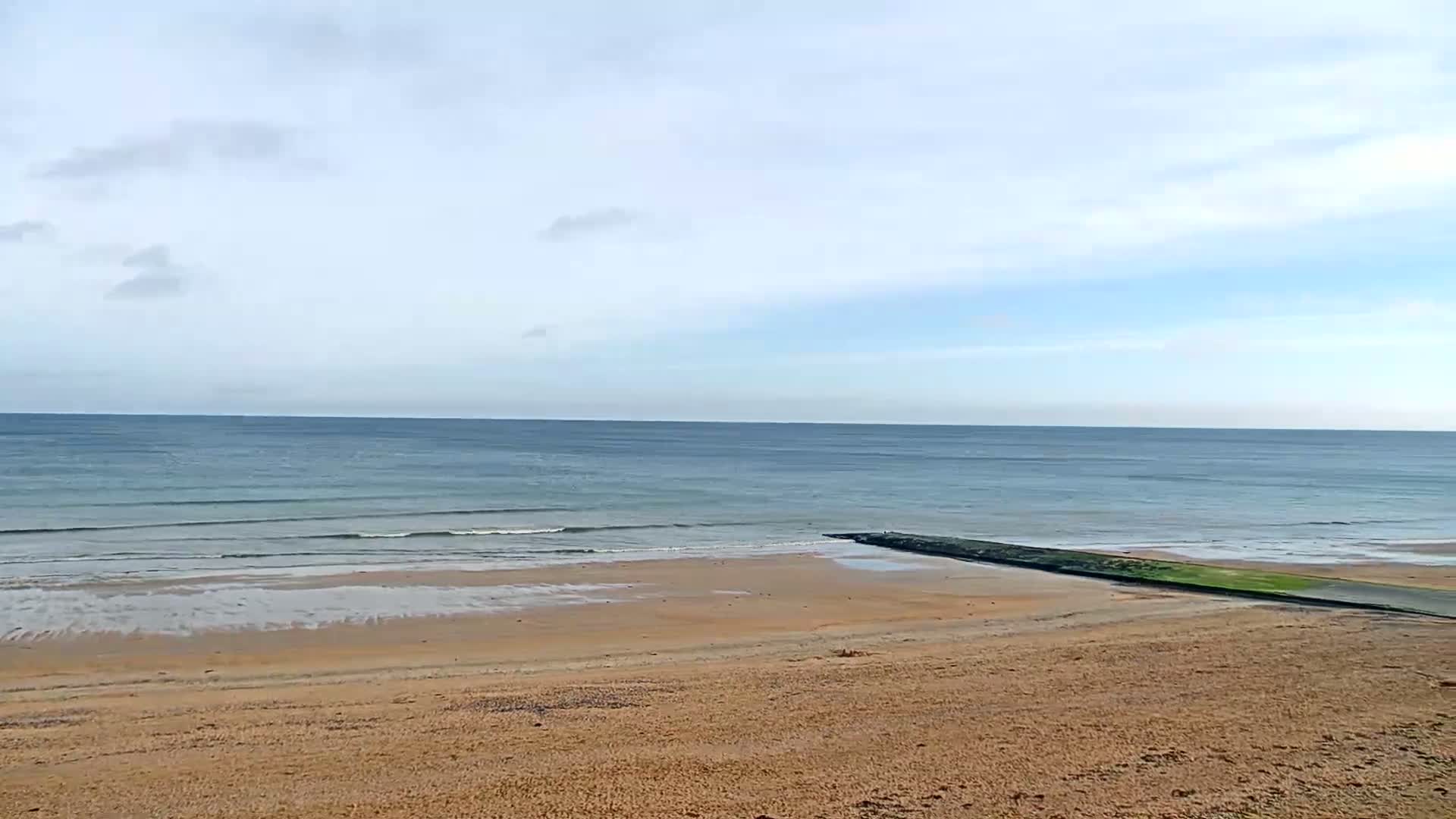 A sandy beach slopes down to a calm, blue ocean under a mostly cloudy sky, with a low-lying breakwater extending from the shore into the water.