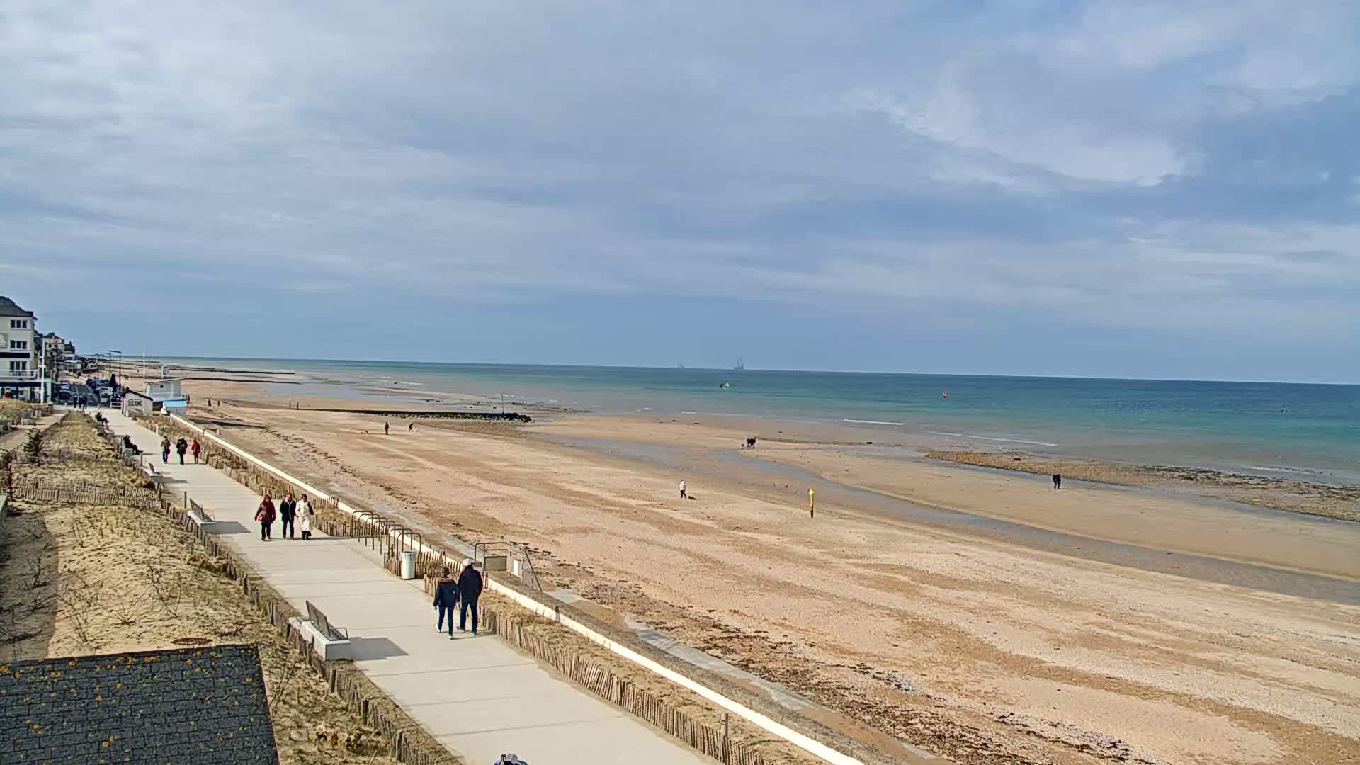 A sandy beach with a paved walkway and several people walking stretches out to a calm ocean under a partly cloudy sky.