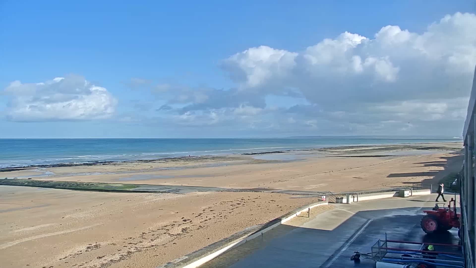 A sandy beach stretches to a calm ocean under a partly cloudy blue sky, with a tractor parked nearby.