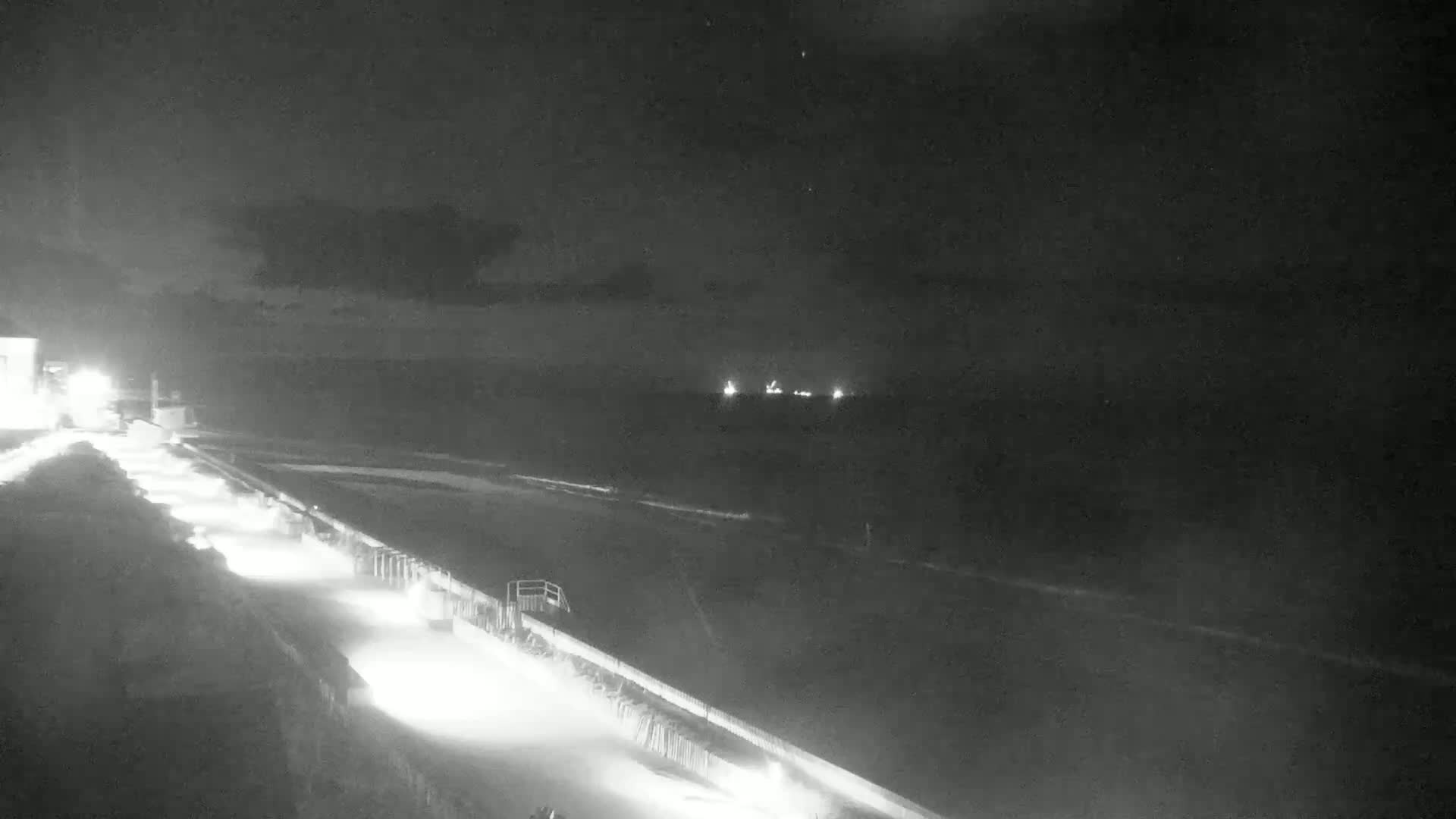 A nighttime, low-light view shows a dark beach with gently lapping waves and a paved walkway lit by lamps, leading to a dark body of water with faint lights on the horizon.
