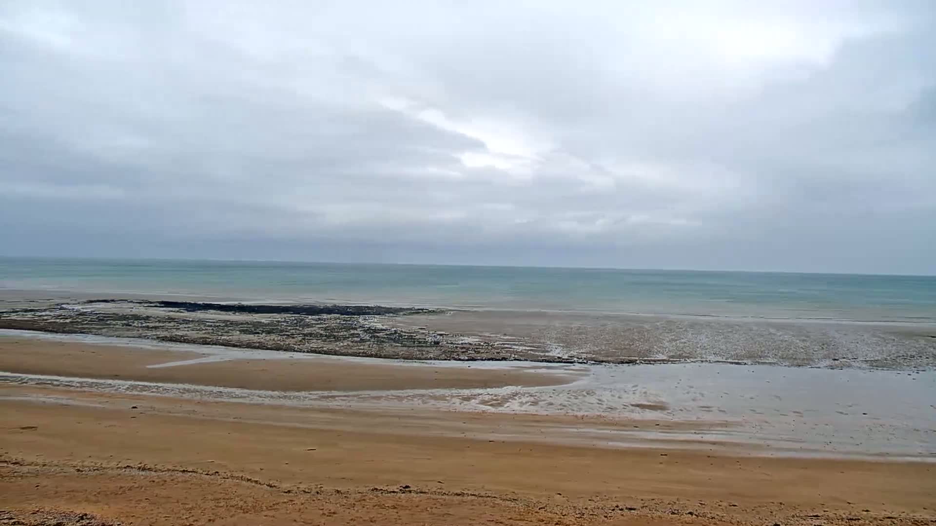 A sandy beach stretches towards a calm, light teal ocean under an overcast sky.