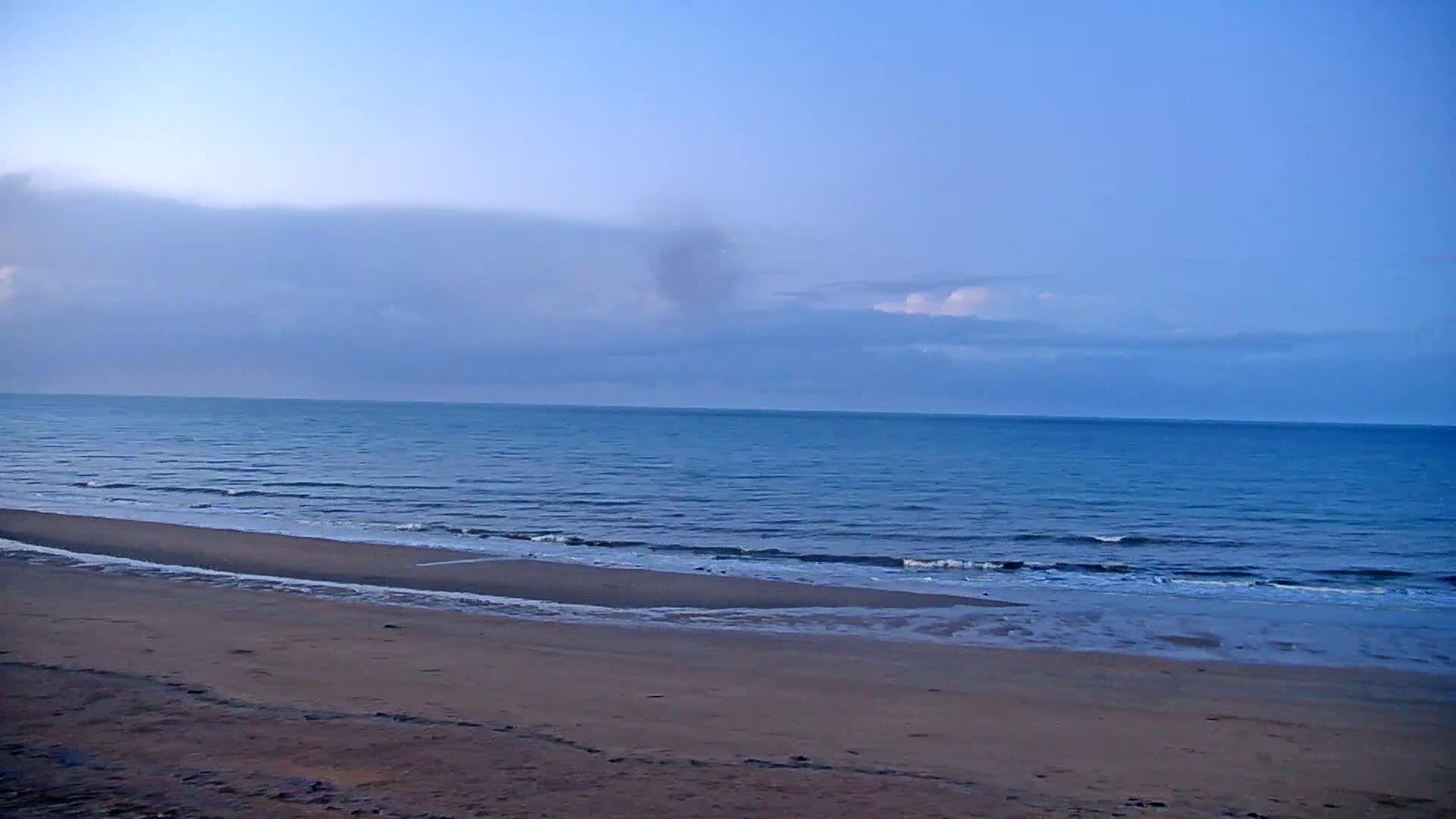 A sandy beach meets a calm, blue ocean under a mostly cloudy, pale blue sky.