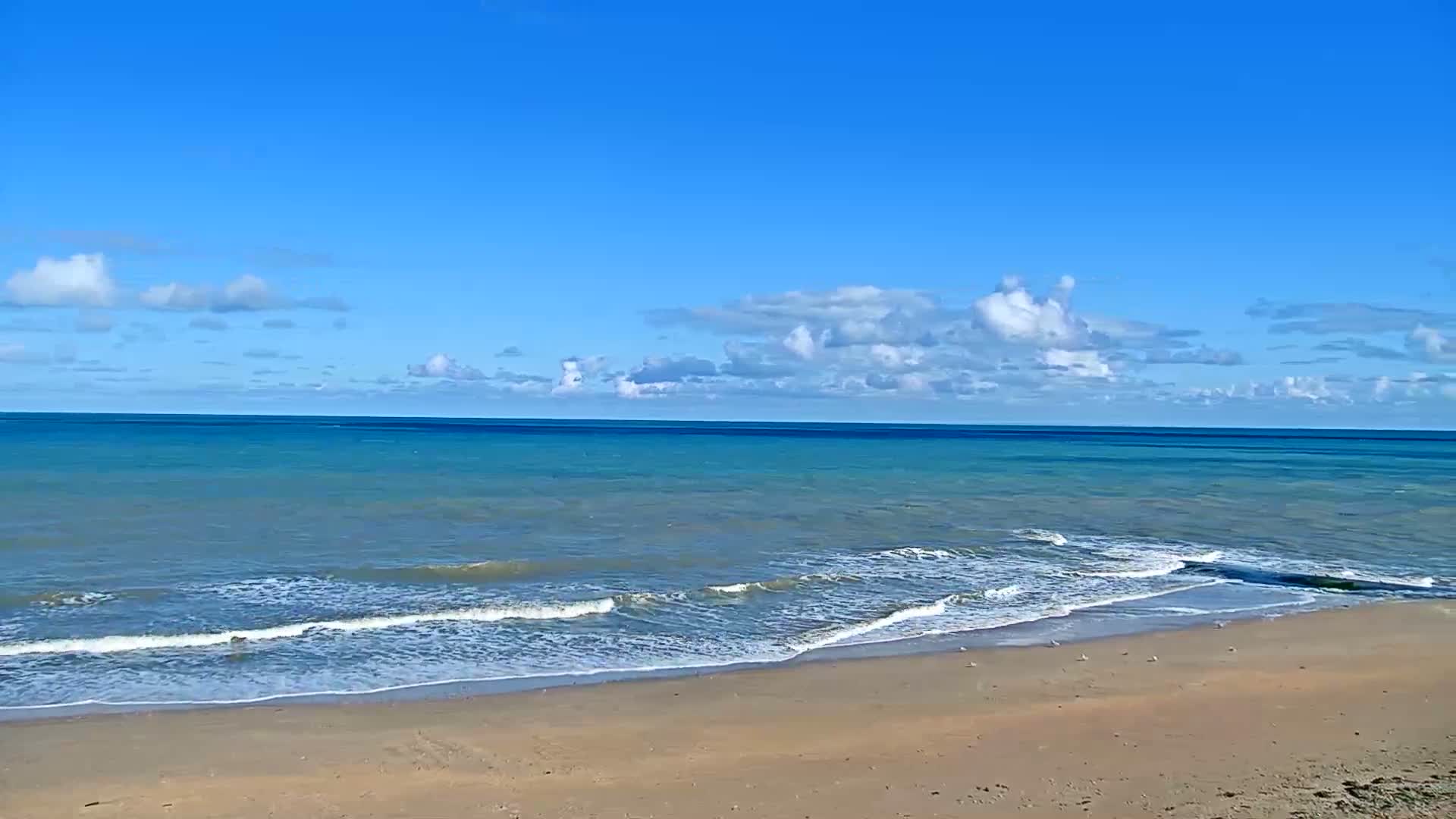 A sandy beach meets a calm, turquoise ocean under a clear blue sky with scattered white clouds.