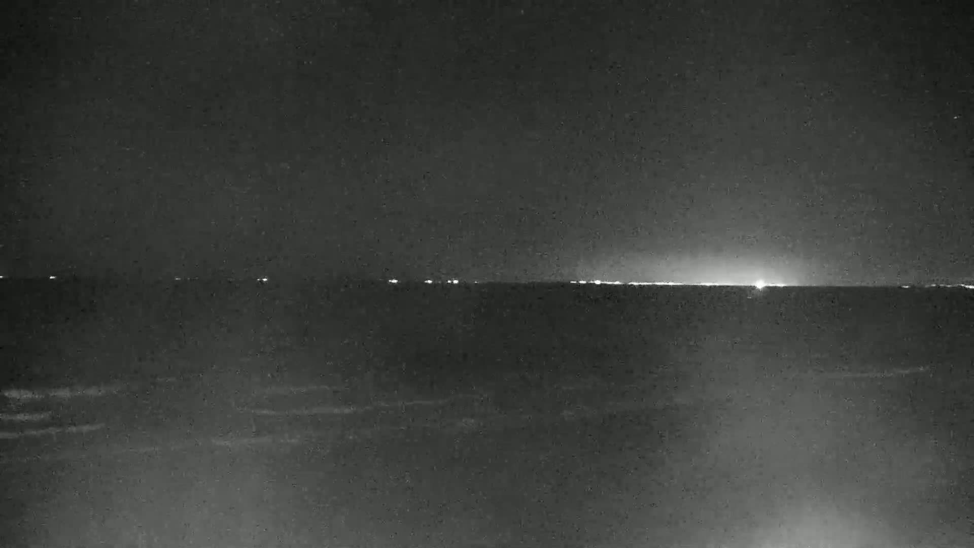 A dark nighttime scene shows a calm body of water with a faint horizon line of distant lights under a clear, starlit sky.