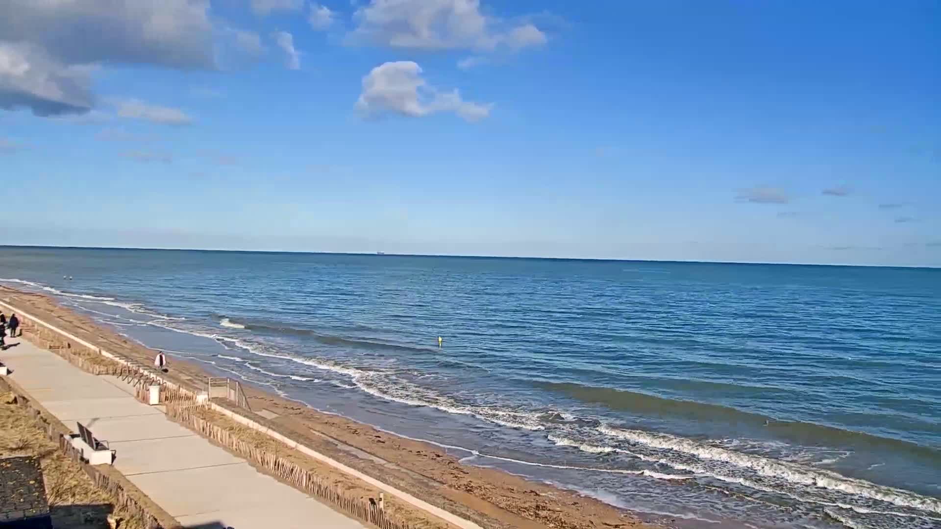 A paved walkway runs alongside a sandy beach and a calm, blue ocean under a mostly sunny sky.