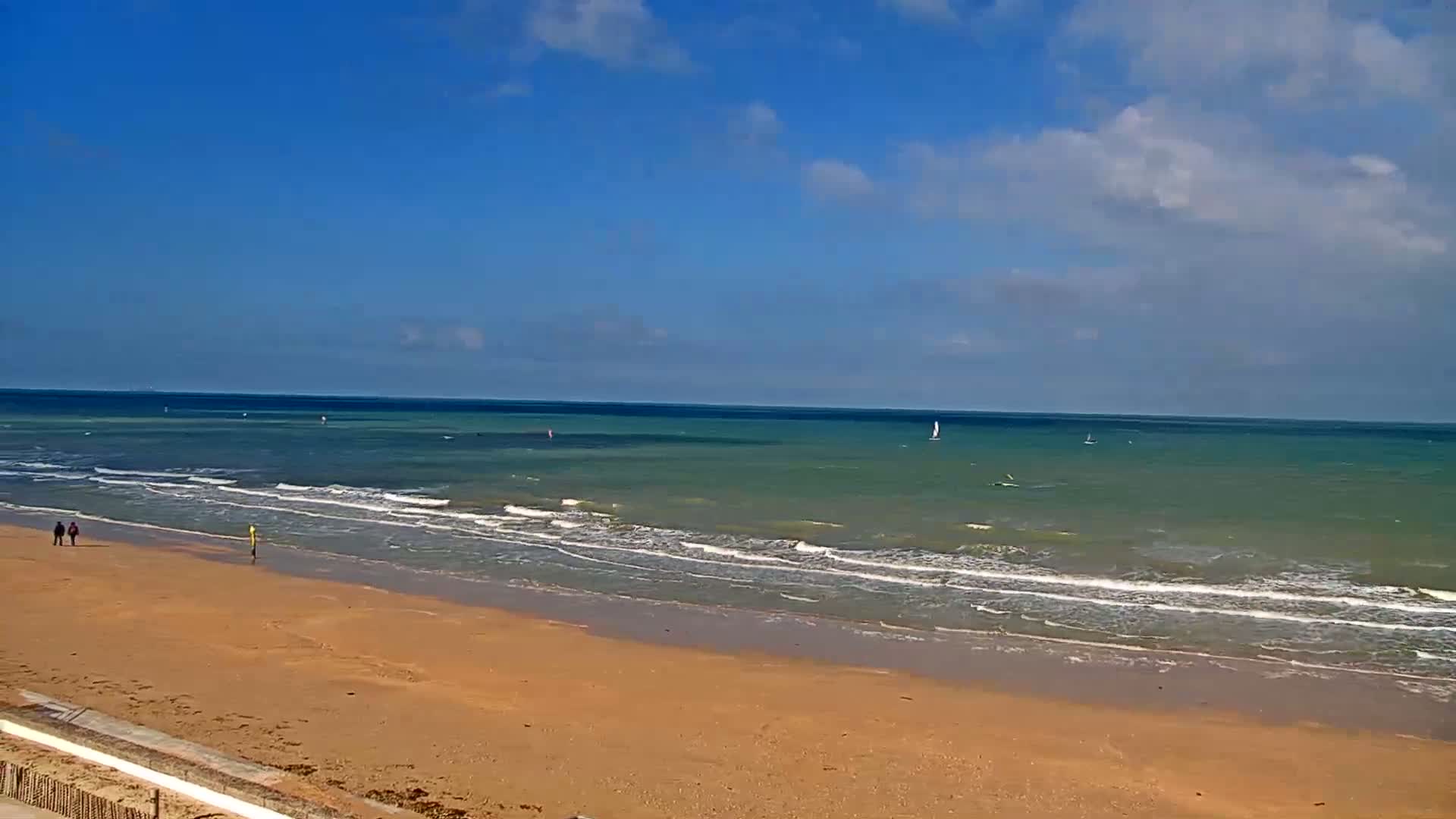 A sandy beach with gently rolling waves under a partly cloudy blue sky, with a few people walking along the shore and sailboats visible on the horizon.