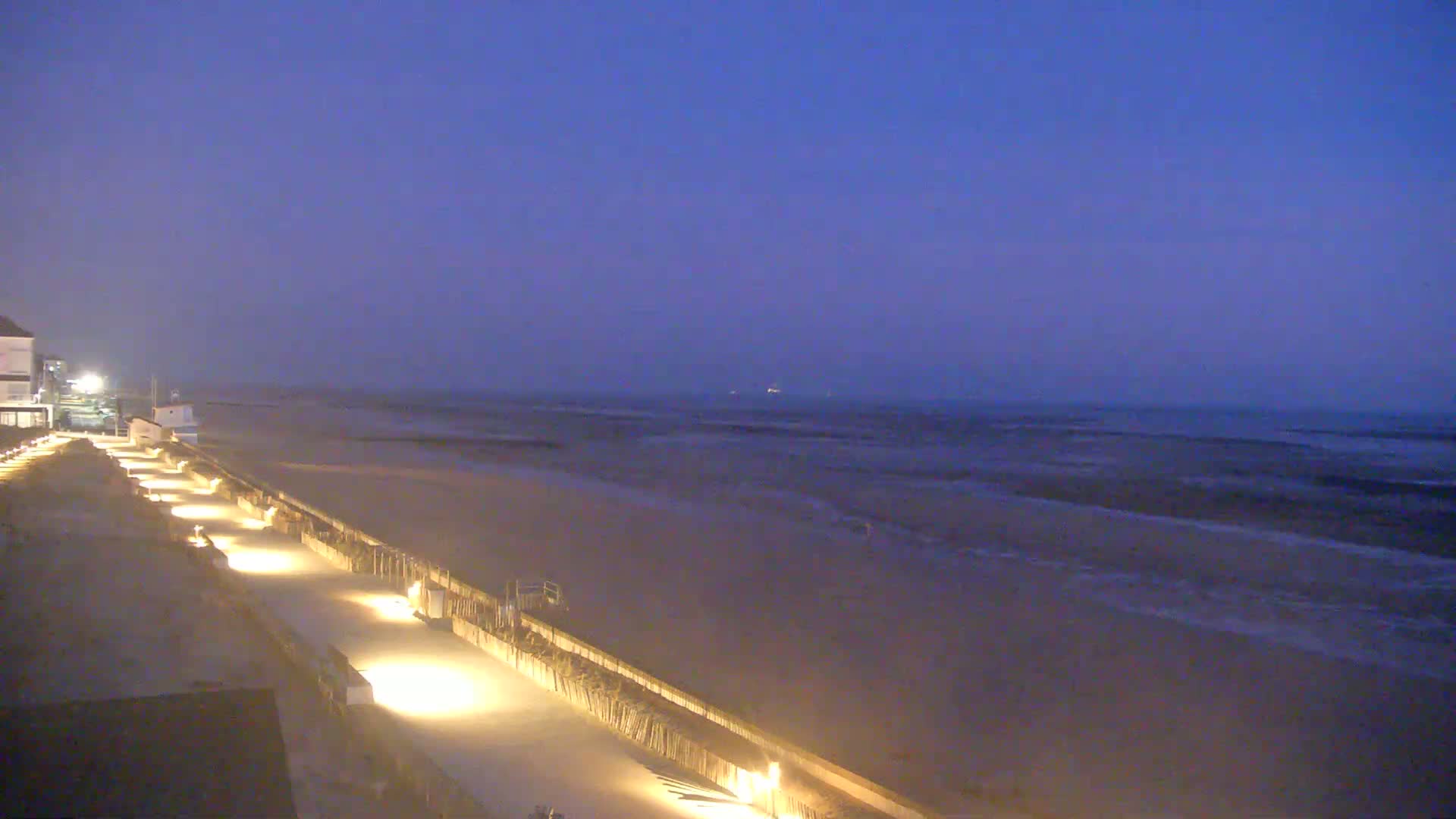 A dimly lit beachfront walkway stretches alongside a sandy beach and a dark, gently waving sea under a twilight sky.