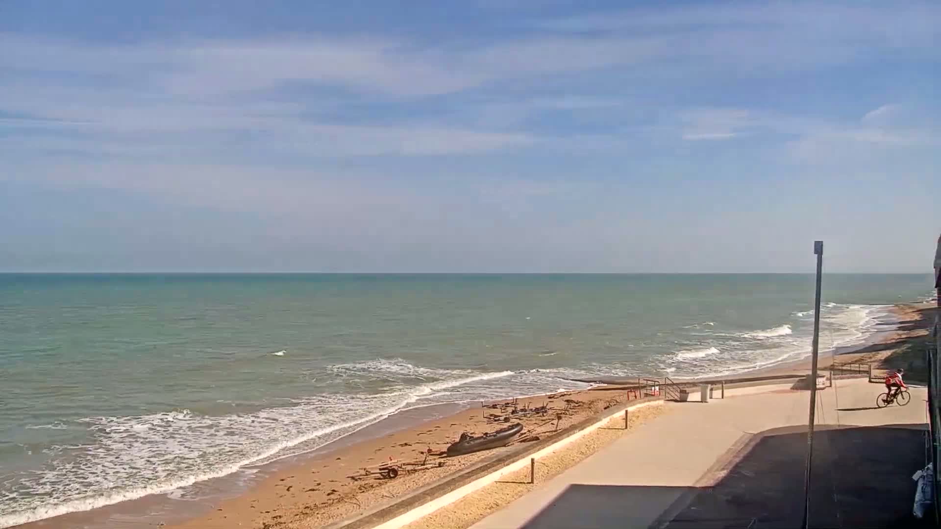 A sandy beach with gentle waves under a mostly sunny sky has a person biking along a paved path above it, with a small inflatable boat and other equipment on the sand.