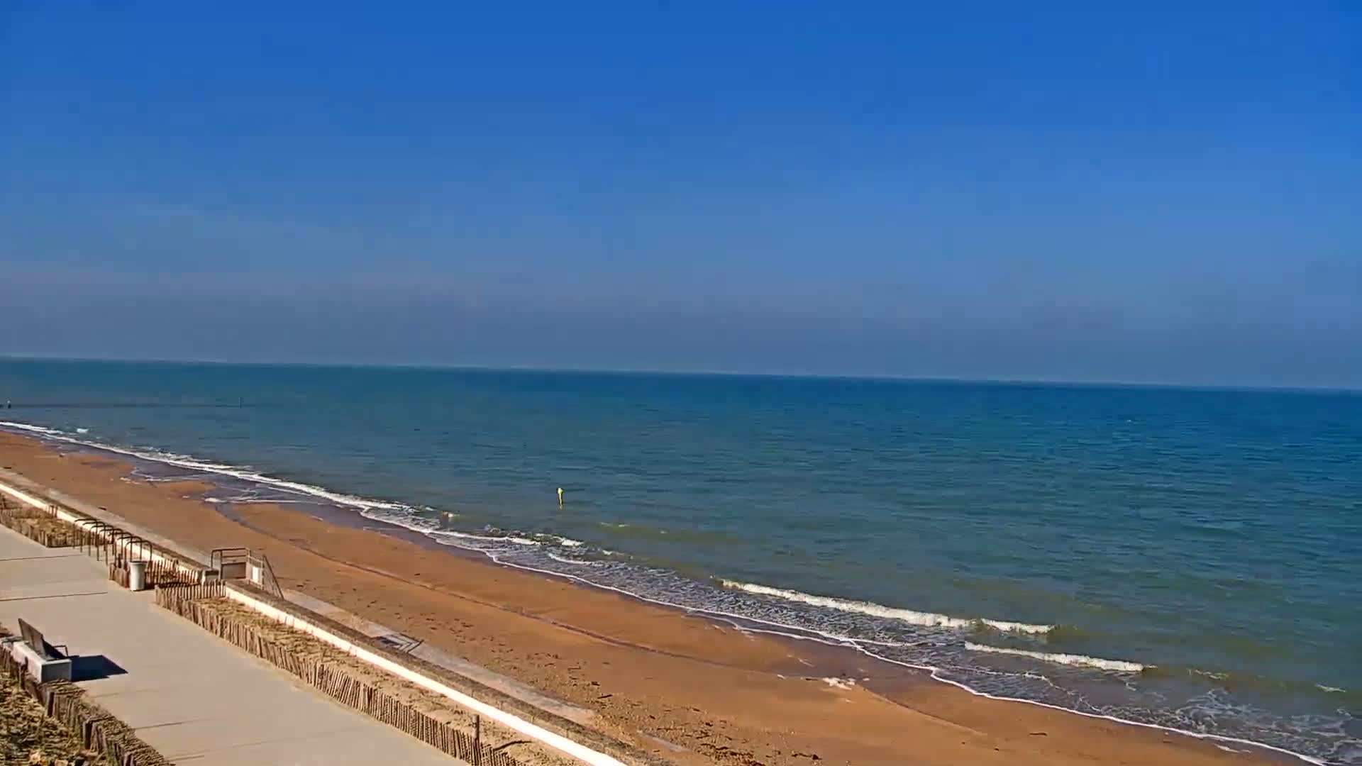 A paved walkway overlooks a sandy beach and calm ocean under a clear blue sky.