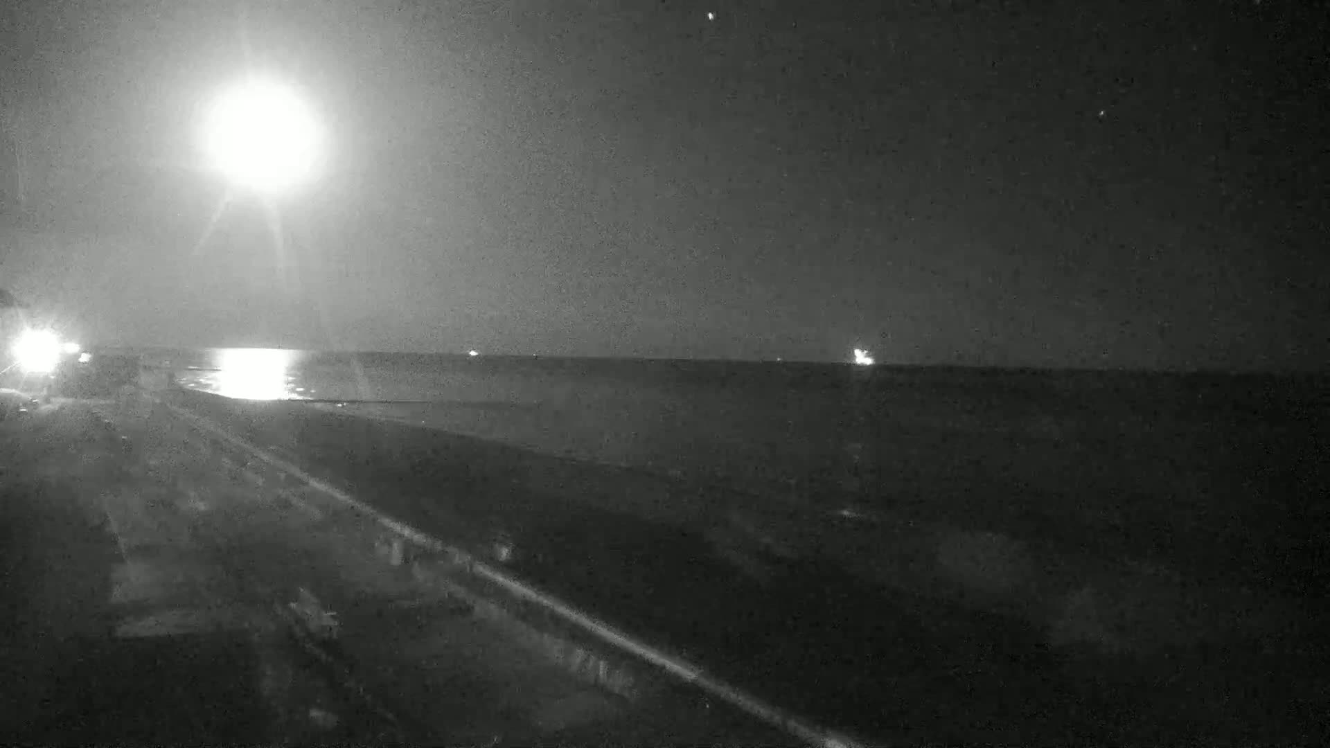 A nighttime view of a dark road next to a calm body of water under a bright moon.
