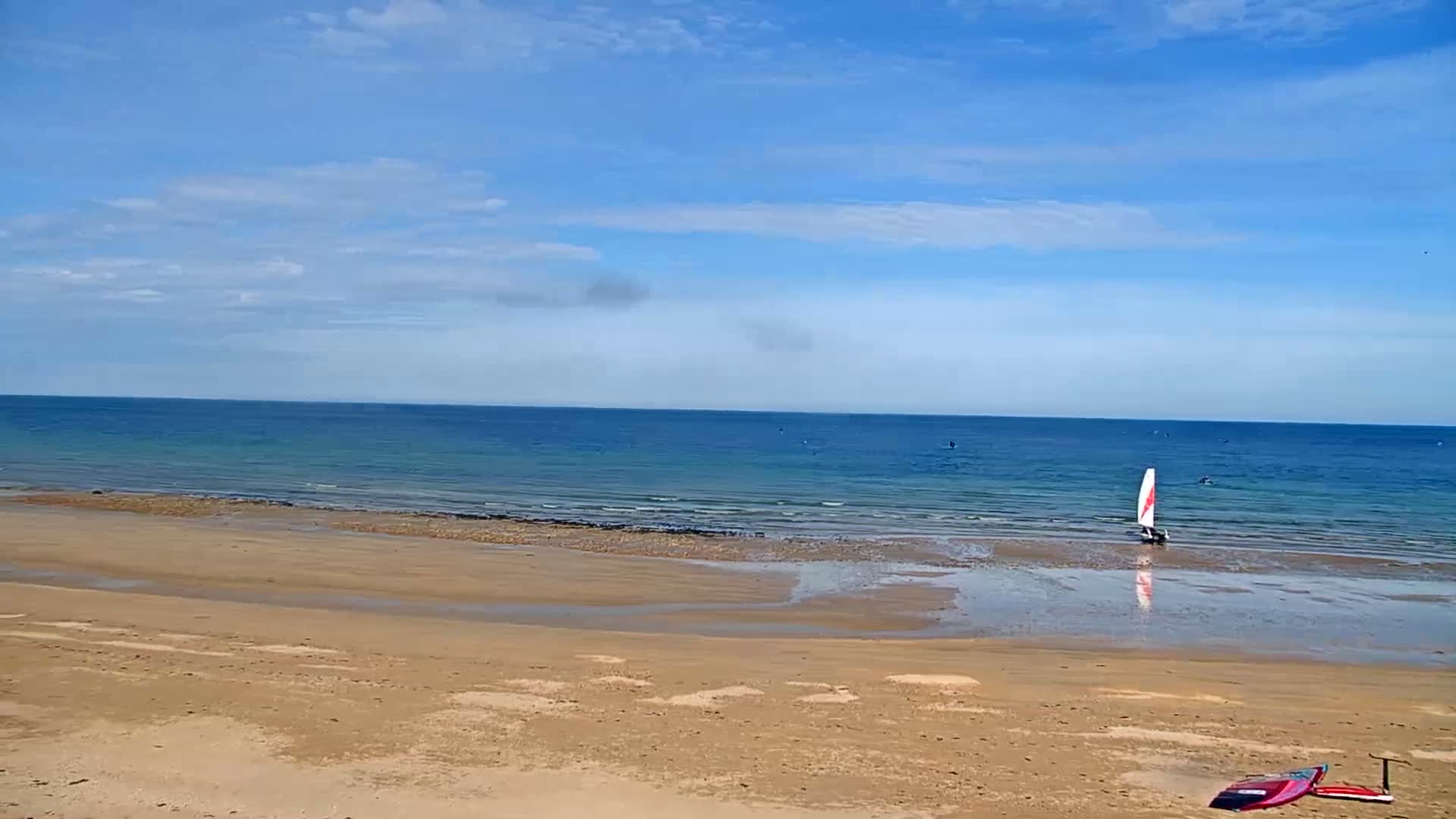 A sandy beach stretches to a calm, blue ocean under a mostly sunny sky, with a small sailboat in the water and a couple of surfboards on the sand.