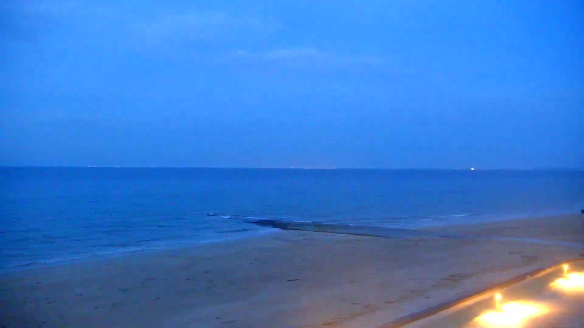 A sandy beach at dusk meets a calm, dark blue ocean under a clear twilight sky.
