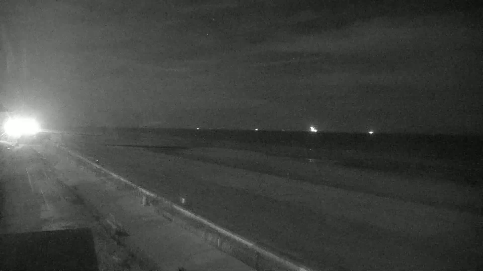 A nighttime view of a long, dark road next to a beach, with faint lights visible on the water in the distance under a mostly clear sky.