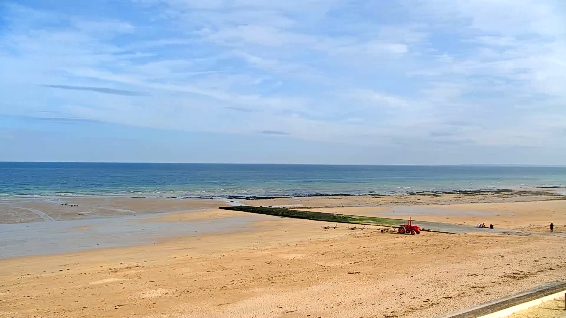 A sandy beach stretches toward a calm ocean under a mostly clear, light blue sky, with a red tractor parked near a paved path leading from the beach to the water.