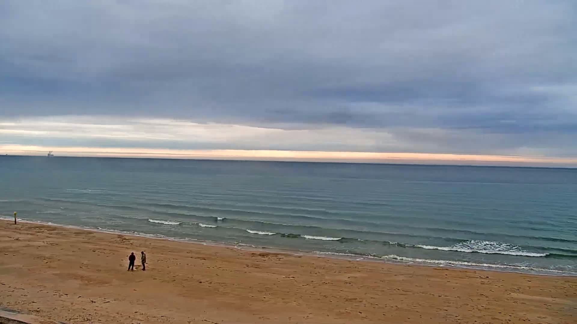 Two people stand on a sandy beach under a cloudy sky, gazing out at a calm sea with small waves.