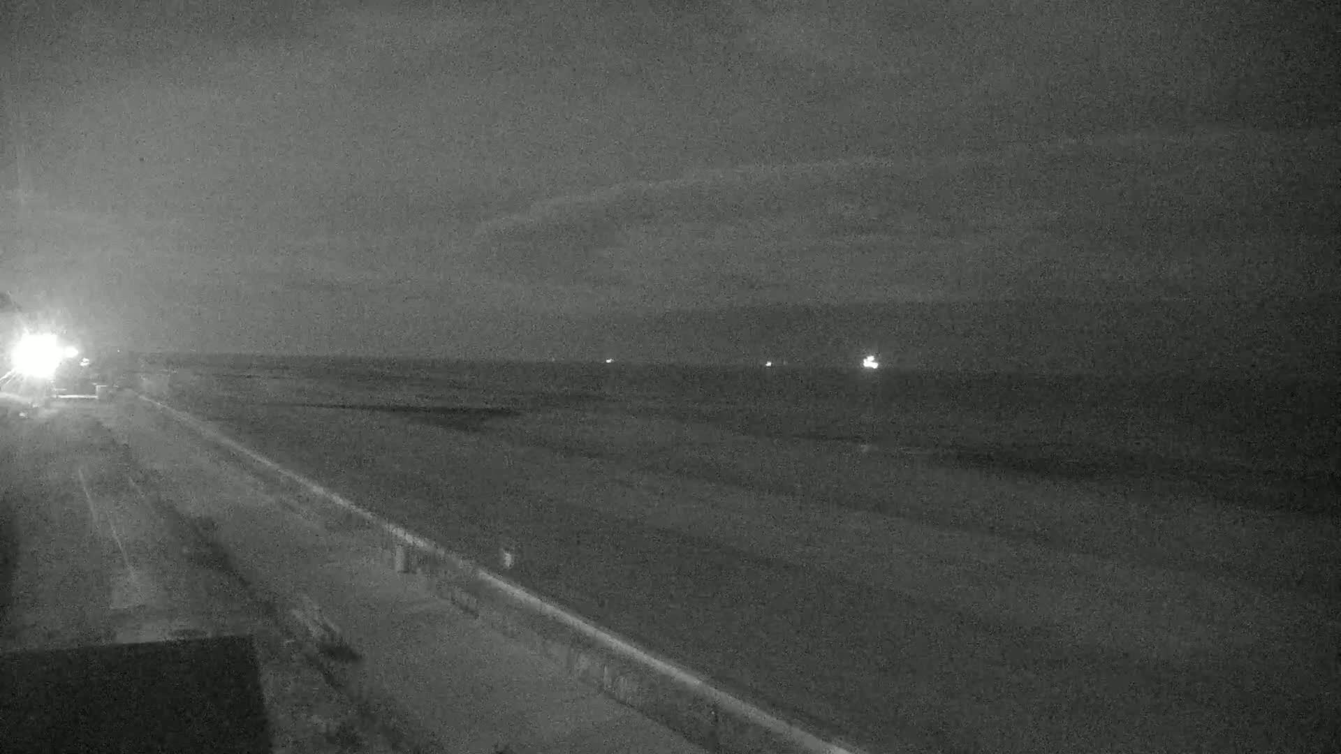 A dark, nighttime view of a beach and adjacent road, with faint lights visible on the water in the distance under a cloudy sky.