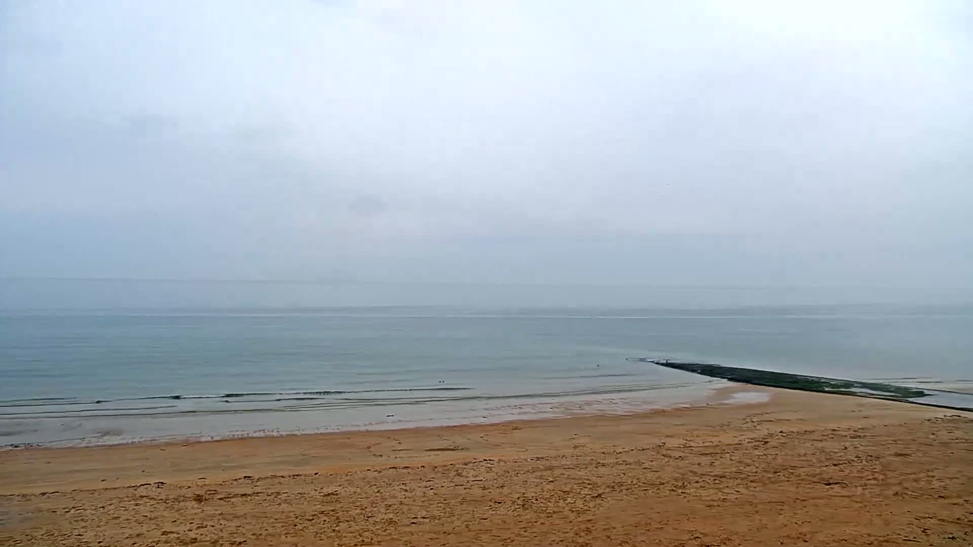 A long, dark structure extends from the shore into a calm, gray sea under an overcast sky.
