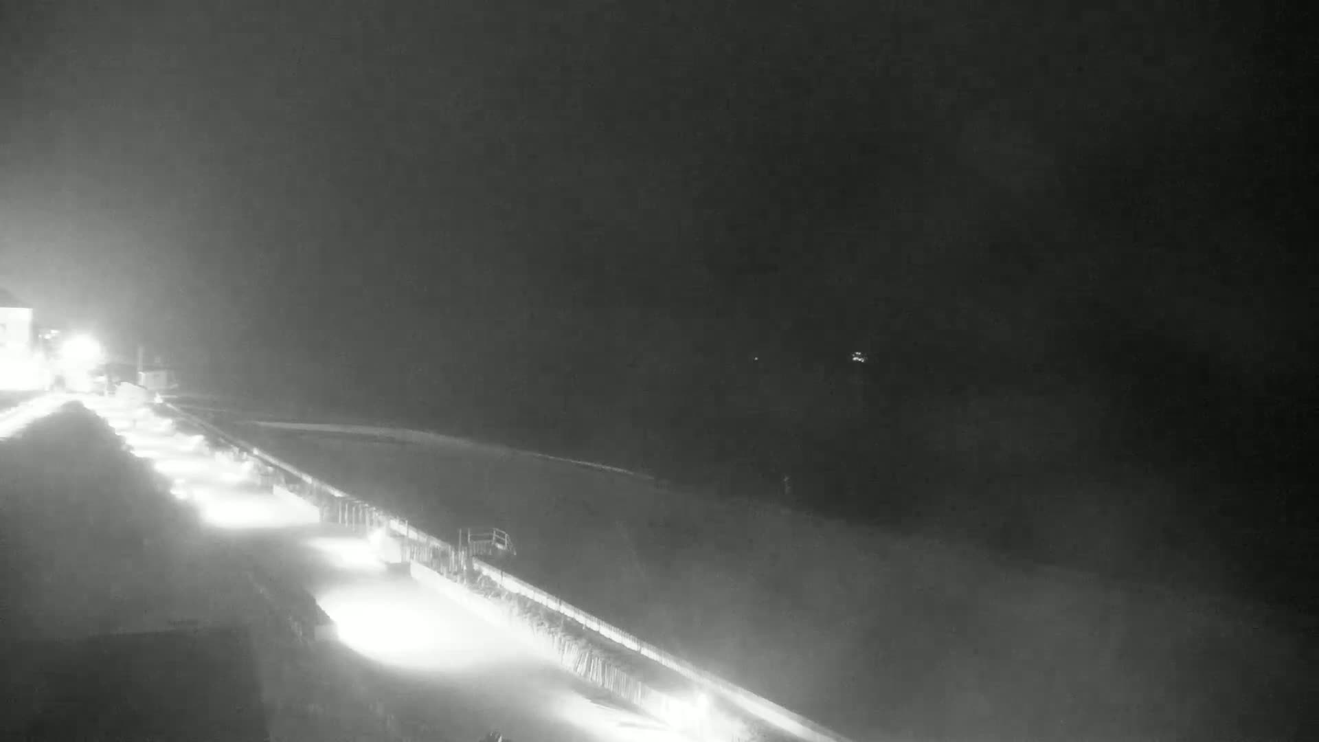 A snow-covered road at night is lit by headlights and streetlights, with a hazy or foggy atmosphere.