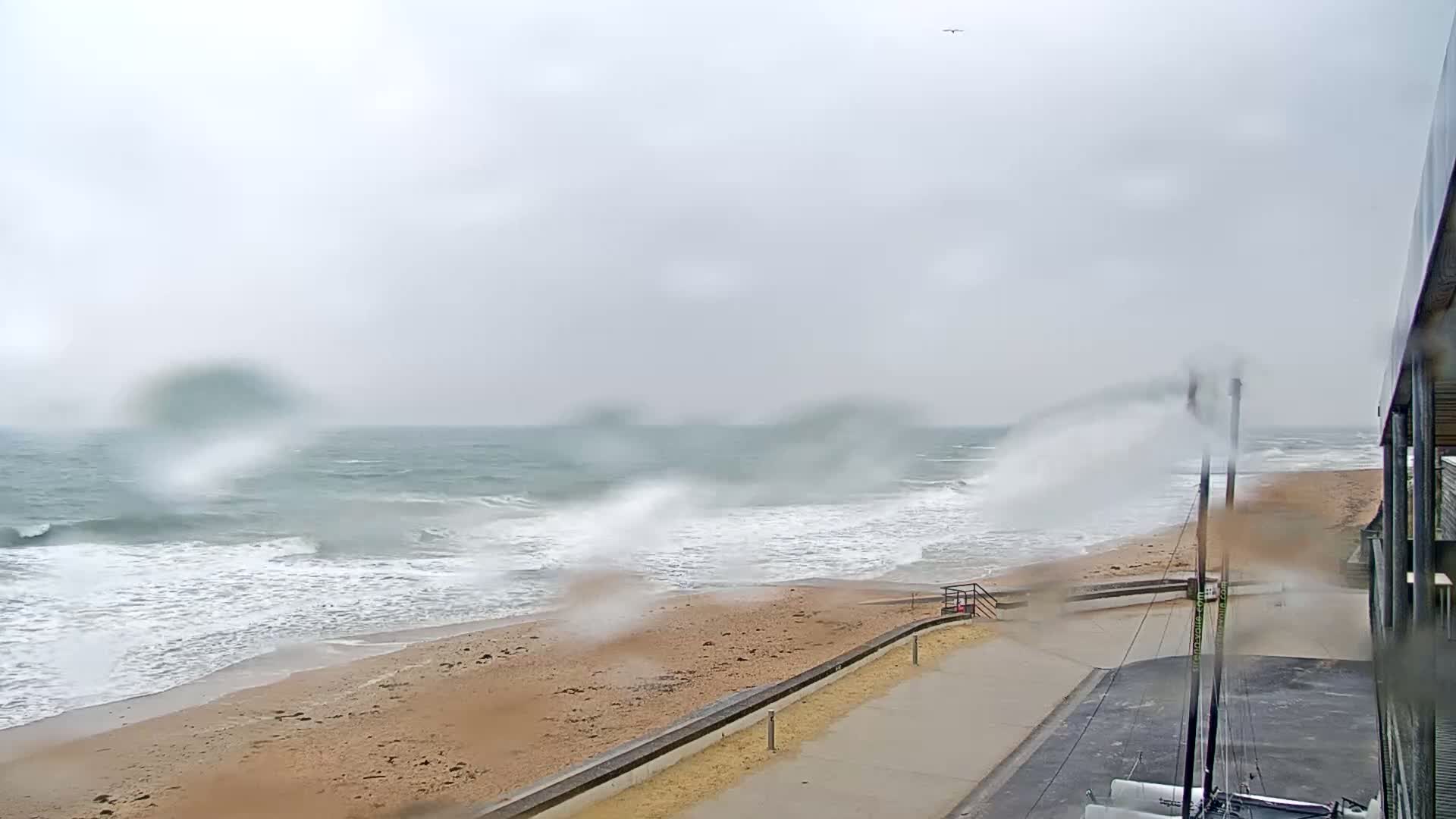 A rain-streaked view shows a beach with choppy waves crashing onto the shore, under an overcast sky.