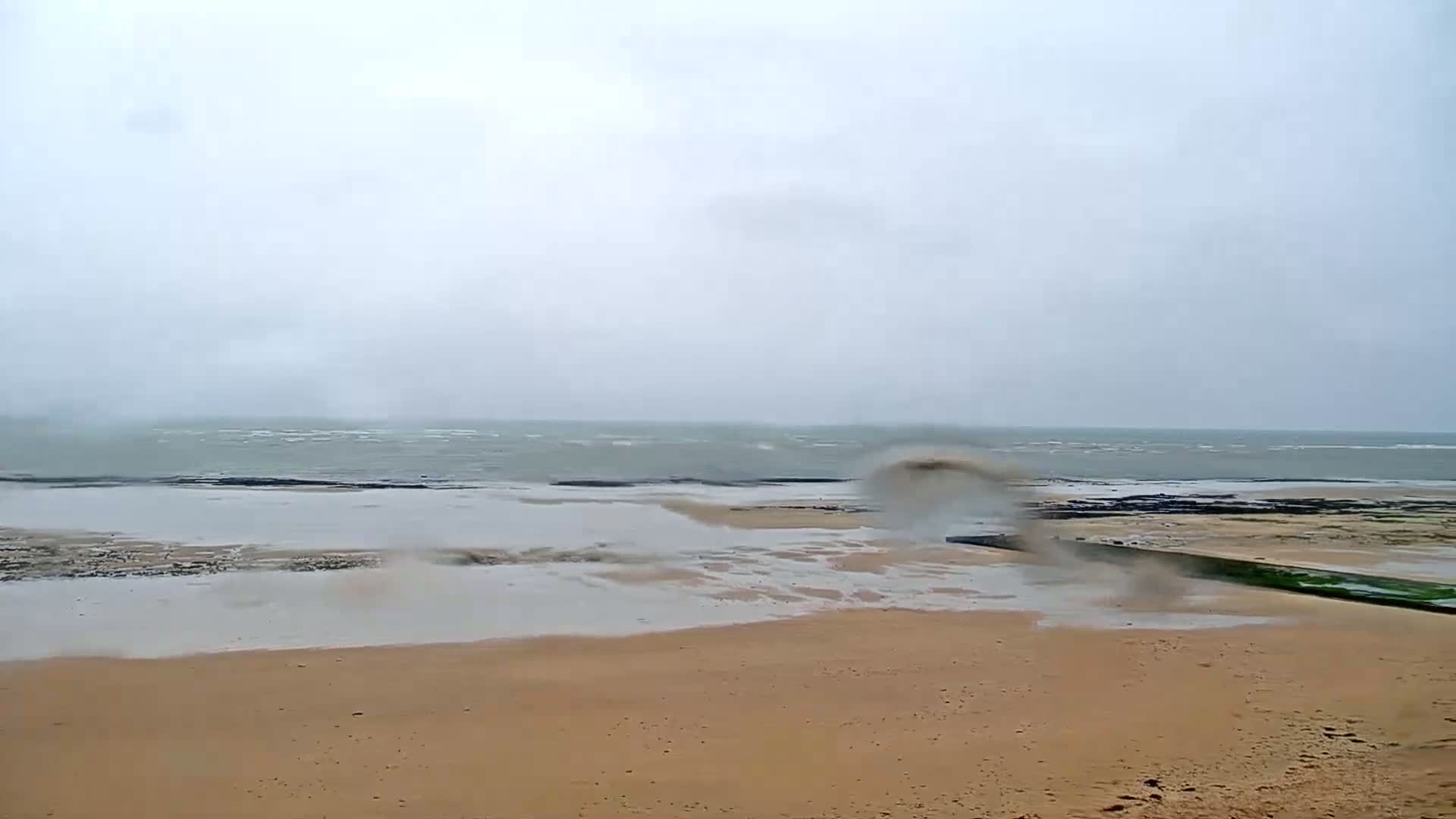 A sandy beach with a low wall is partially submerged by a choppy, grey sea under an overcast sky.