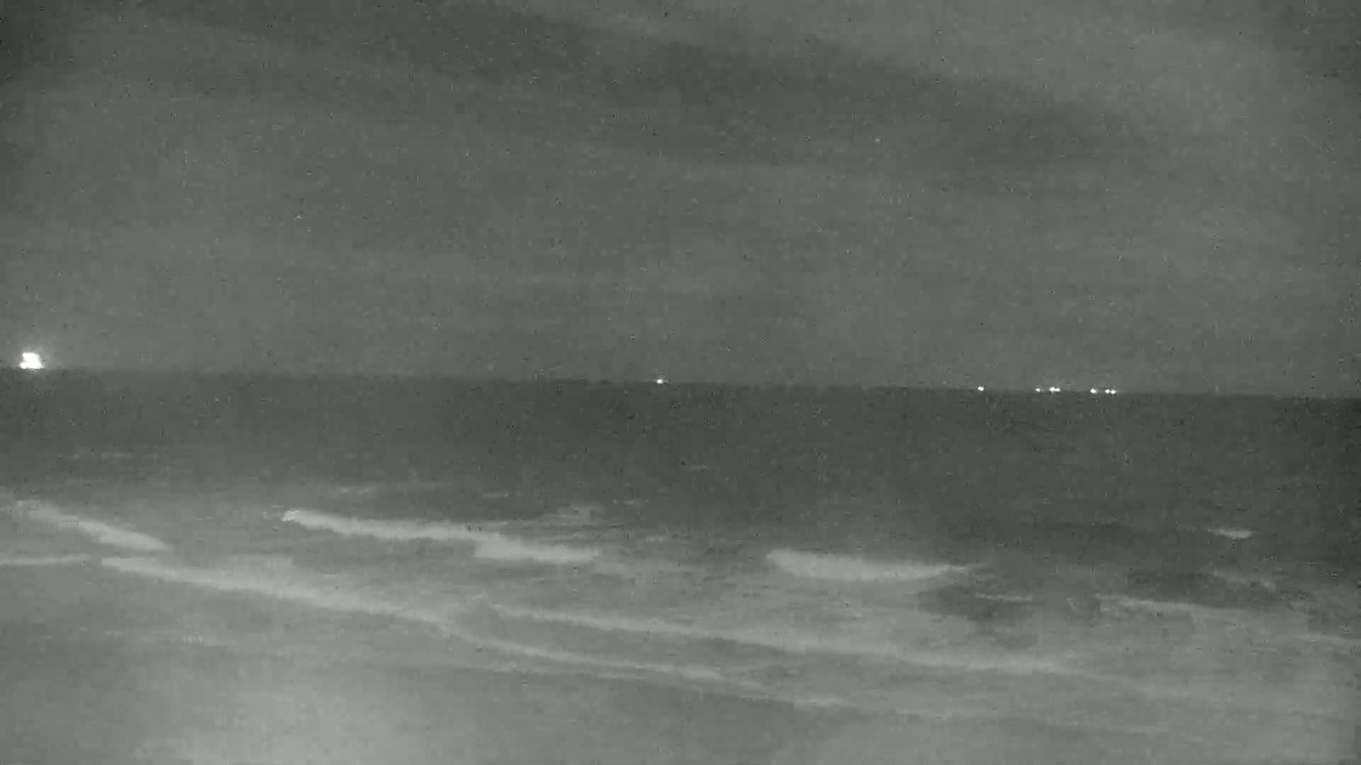 A nighttime, low-light view shows a dark ocean with small waves lapping at a shore under a cloudy sky.
