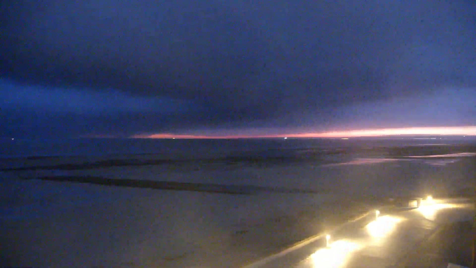 A dark beach at twilight shows a pinkish-orange horizon under a mostly cloudy, dark blue sky, with lights illuminating a path in the foreground.