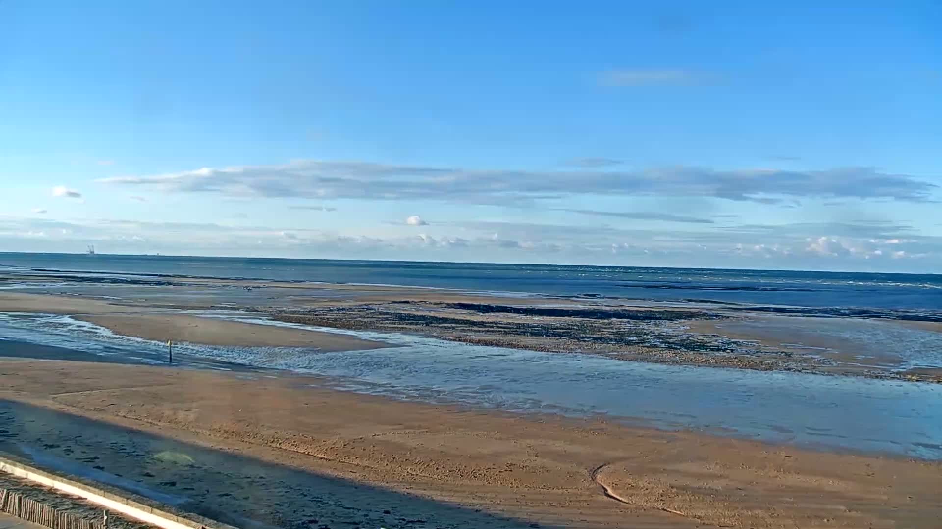 A sandy beach stretches out to a calm sea under a mostly clear blue sky with a few scattered clouds.