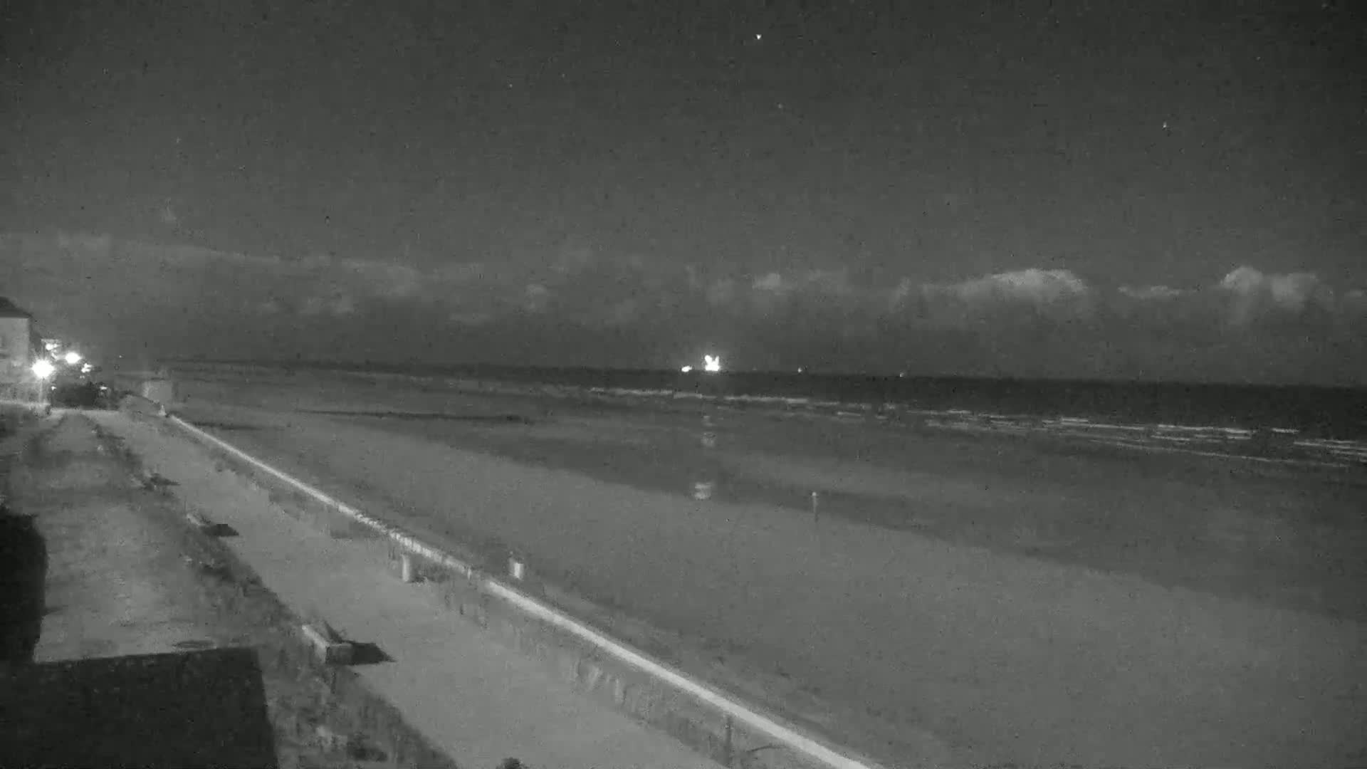 A nighttime beach scene shows a dark ocean with small waves under a mostly clear sky with a few clouds, a wide sandy beach, and a paved walkway next to buildings.