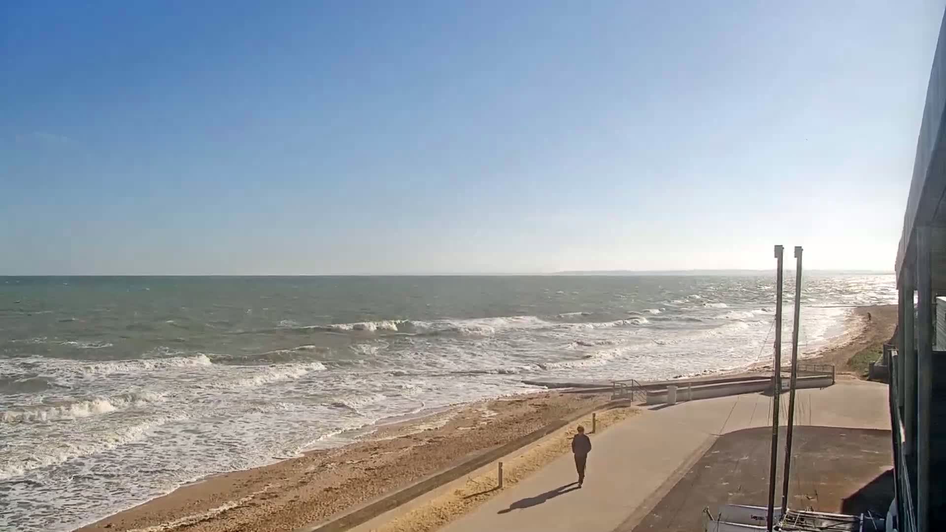 A person walks along a paved path next to a beach with moderate waves under a clear blue sky.