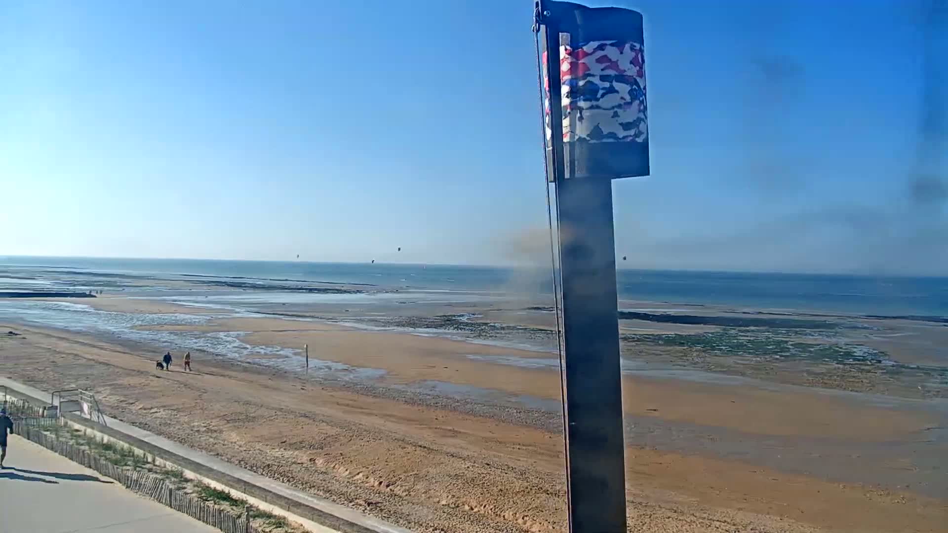A sandy beach stretches to a calm ocean under a clear blue sky, with a few people walking on the beach and some kitesurfing in the distance.