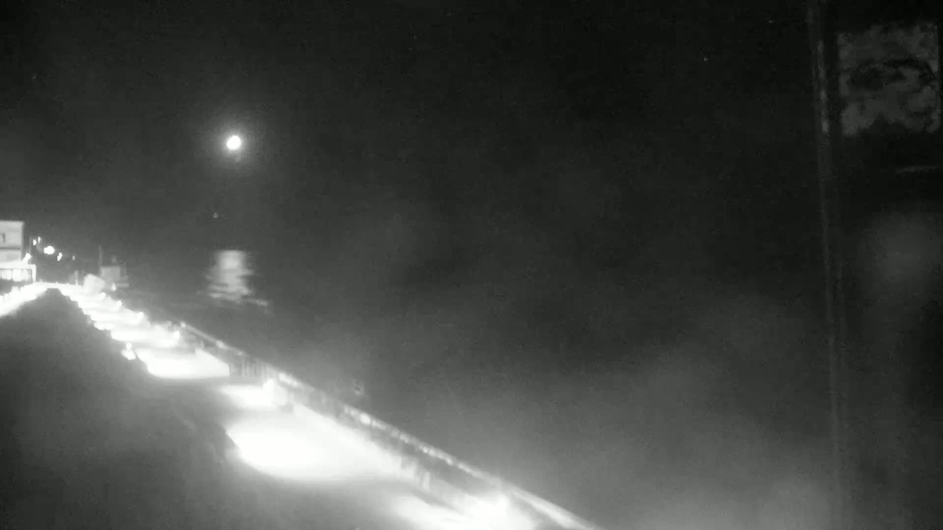 A nighttime, hazy view shows a road with parked cars alongside a body of water under a moonlit sky.