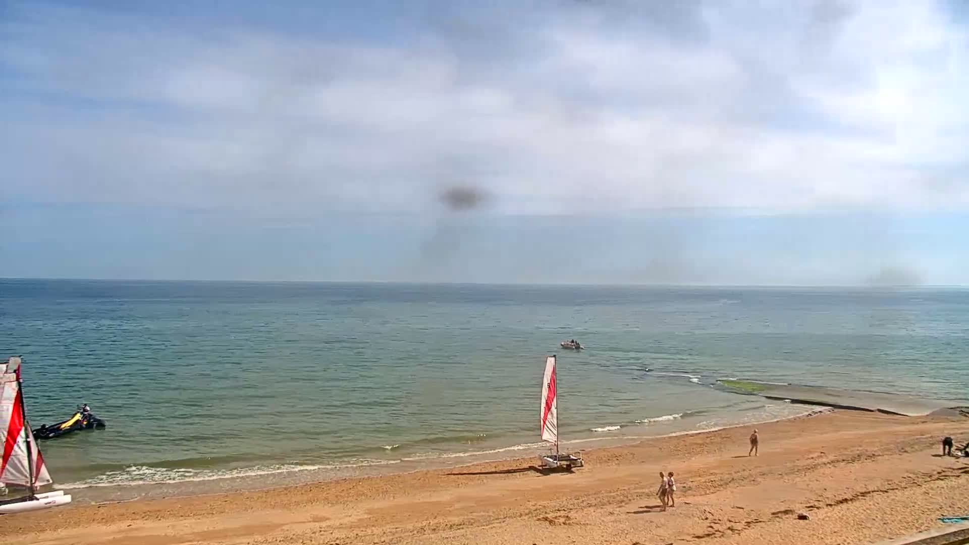 A sandy beach with two small sailboats and several people on it meets a calm, greenish-blue ocean under a partly cloudy sky.