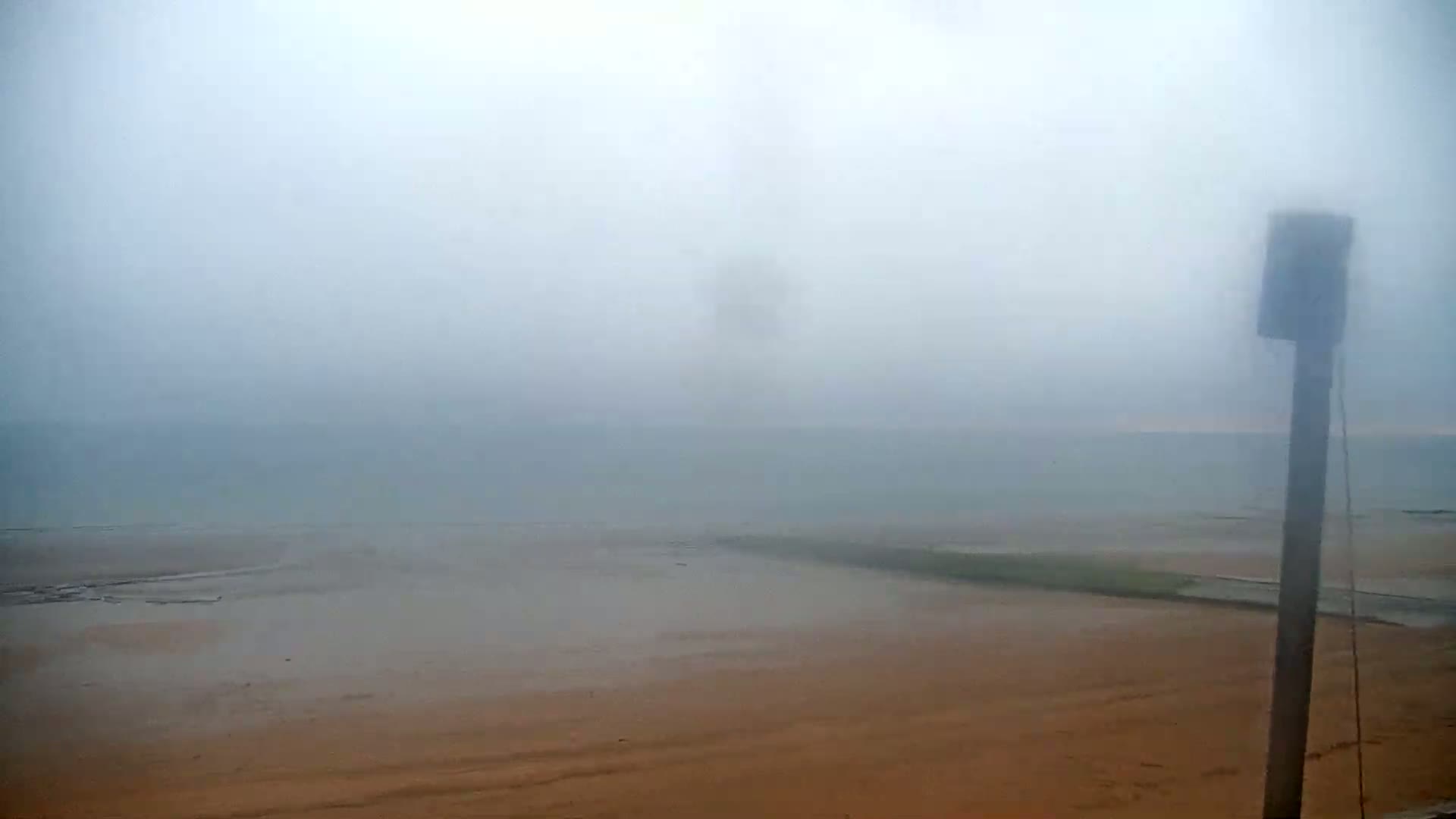 A hazy, foggy day reveals a flat expanse of sandy beach meeting a calm, misty body of water, with a lone post standing in the foreground.