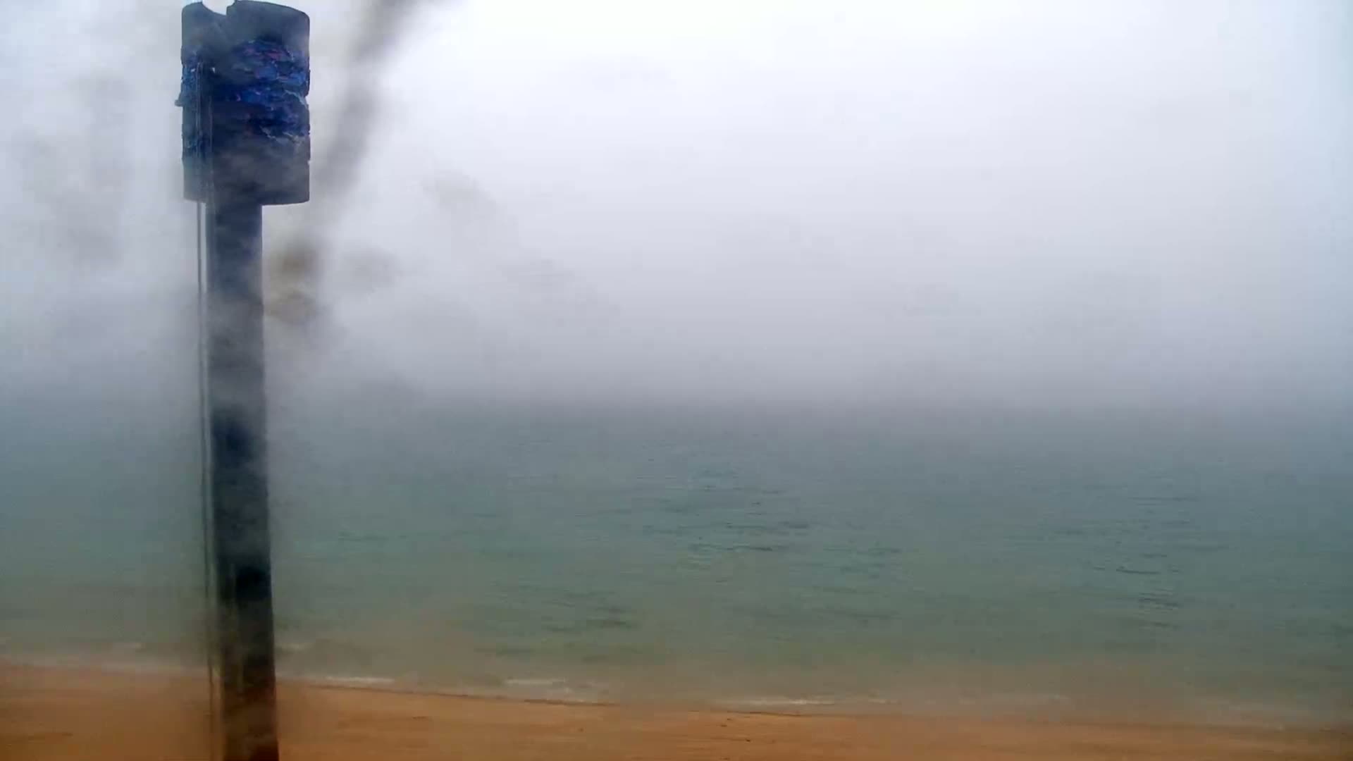 A tall, thin pole with a box-like top stands in front of a foggy ocean and sandy beach.