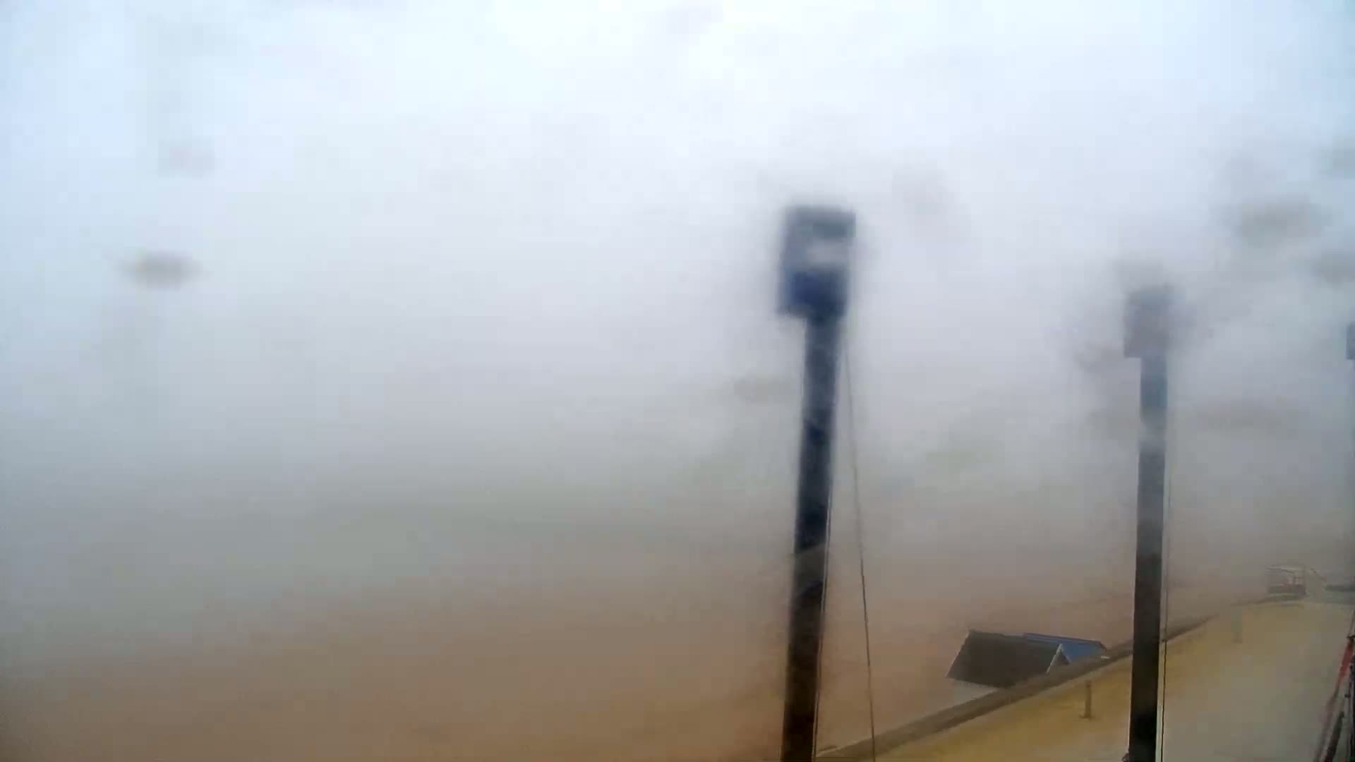 A thick fog obscures the view of a body of water and several tall posts, with a portion of a building visible in the lower right corner.