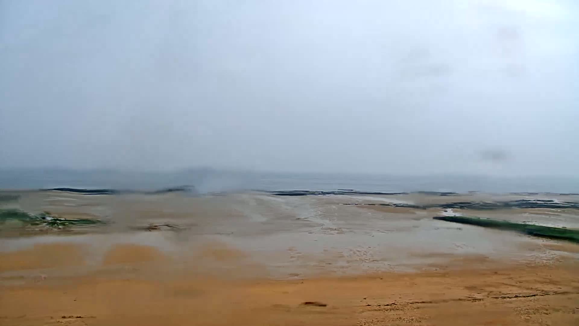 A hazy, rainy day reveals a flat, sandy beach stretching to a calm, gray sea under a muted sky.
