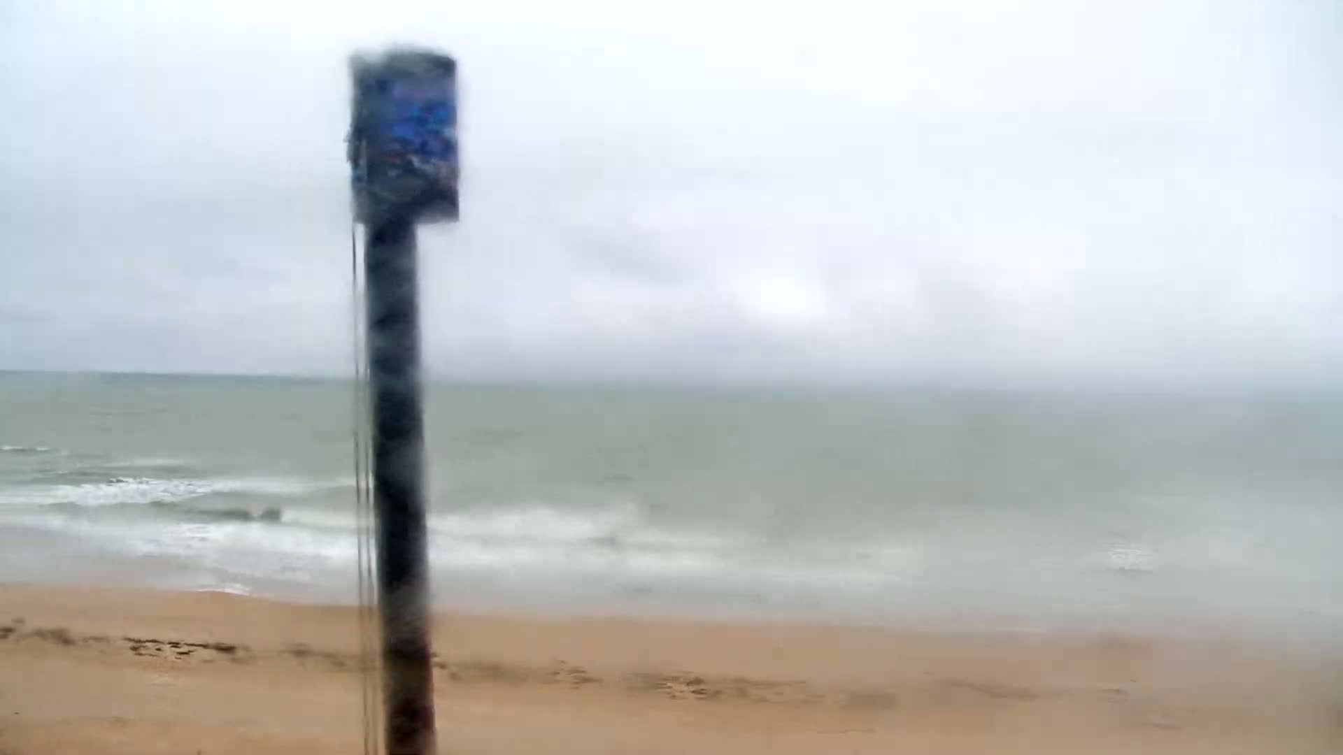 A blurry view through rain-covered glass shows a sandy beach and a gray, wavy ocean under an overcast sky.