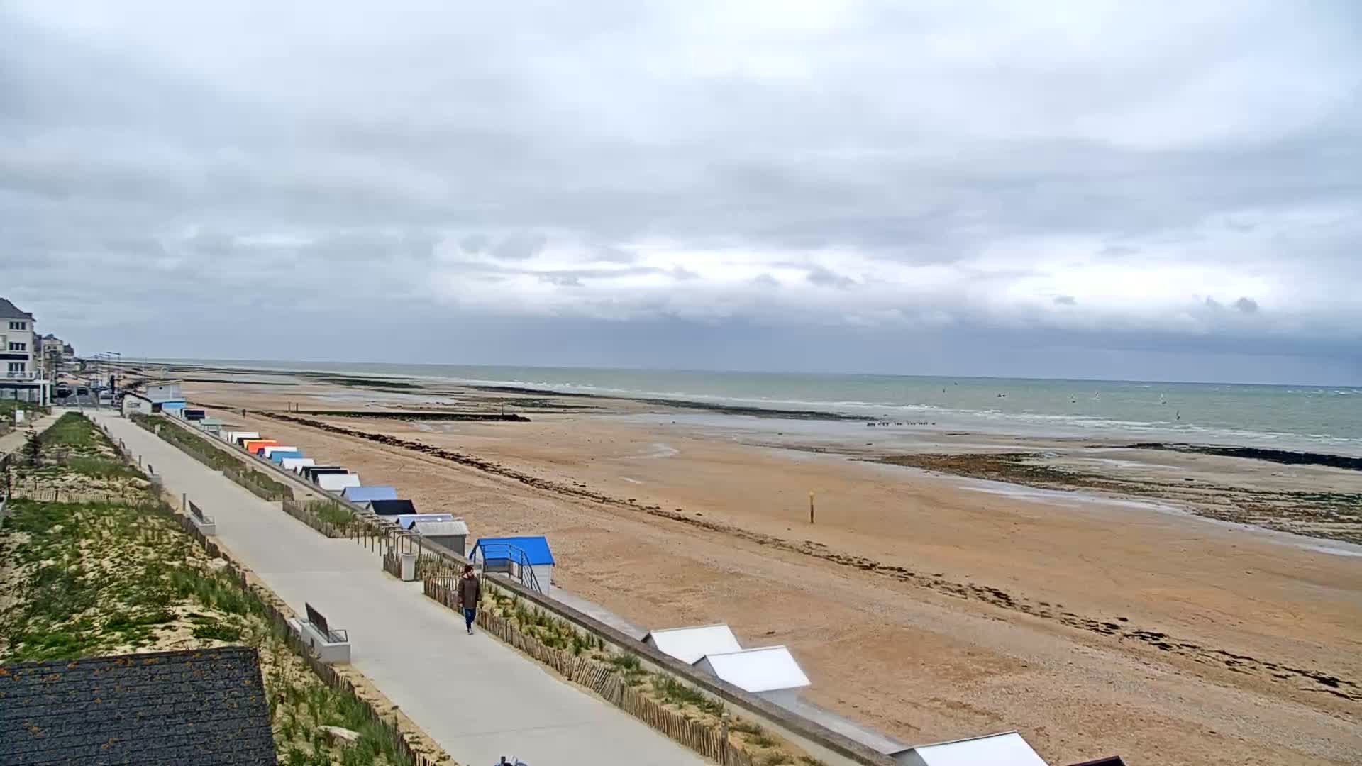 A wide sandy beach stretches to a calm, overcast ocean under a cloudy sky, with a paved walkway lined with colorful beach huts leading from the shore to buildings in the distance.