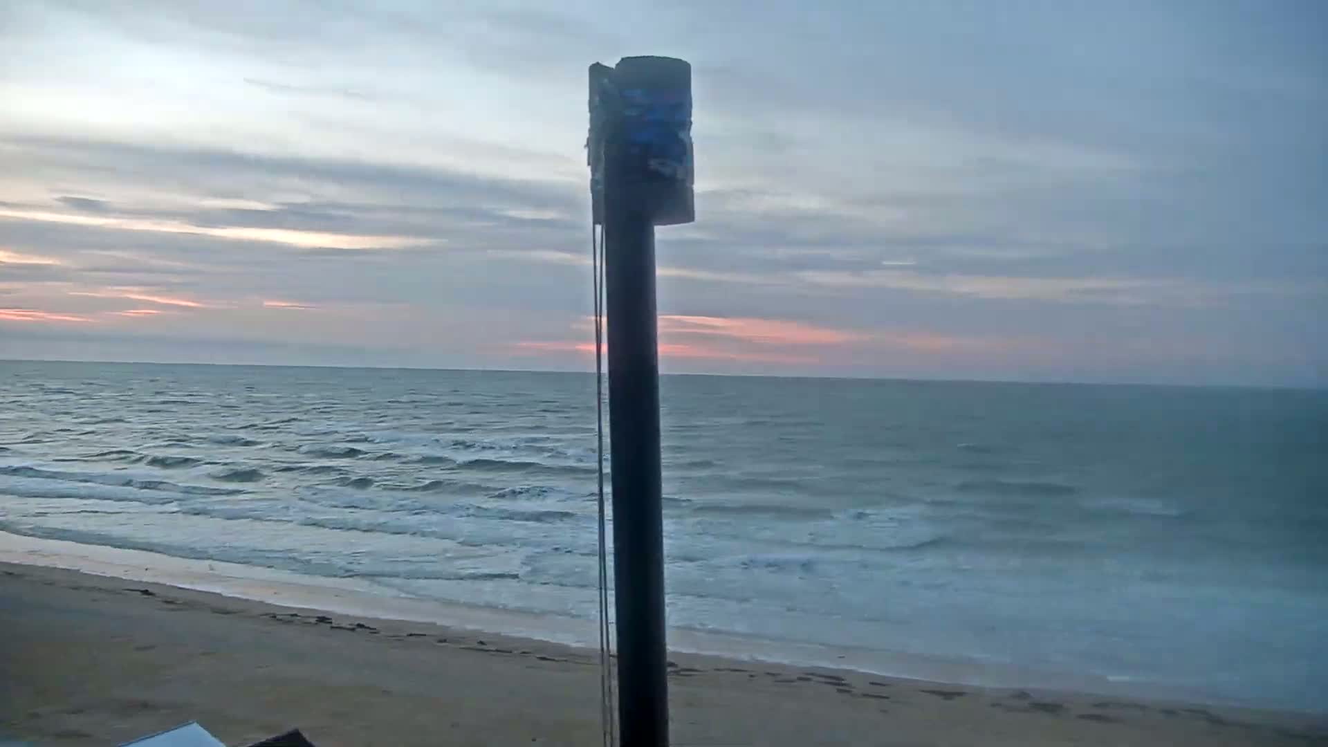 A tall, thin pole stands in the foreground, partially obscuring a view of a calm ocean beach at sunset under a mostly cloudy sky.