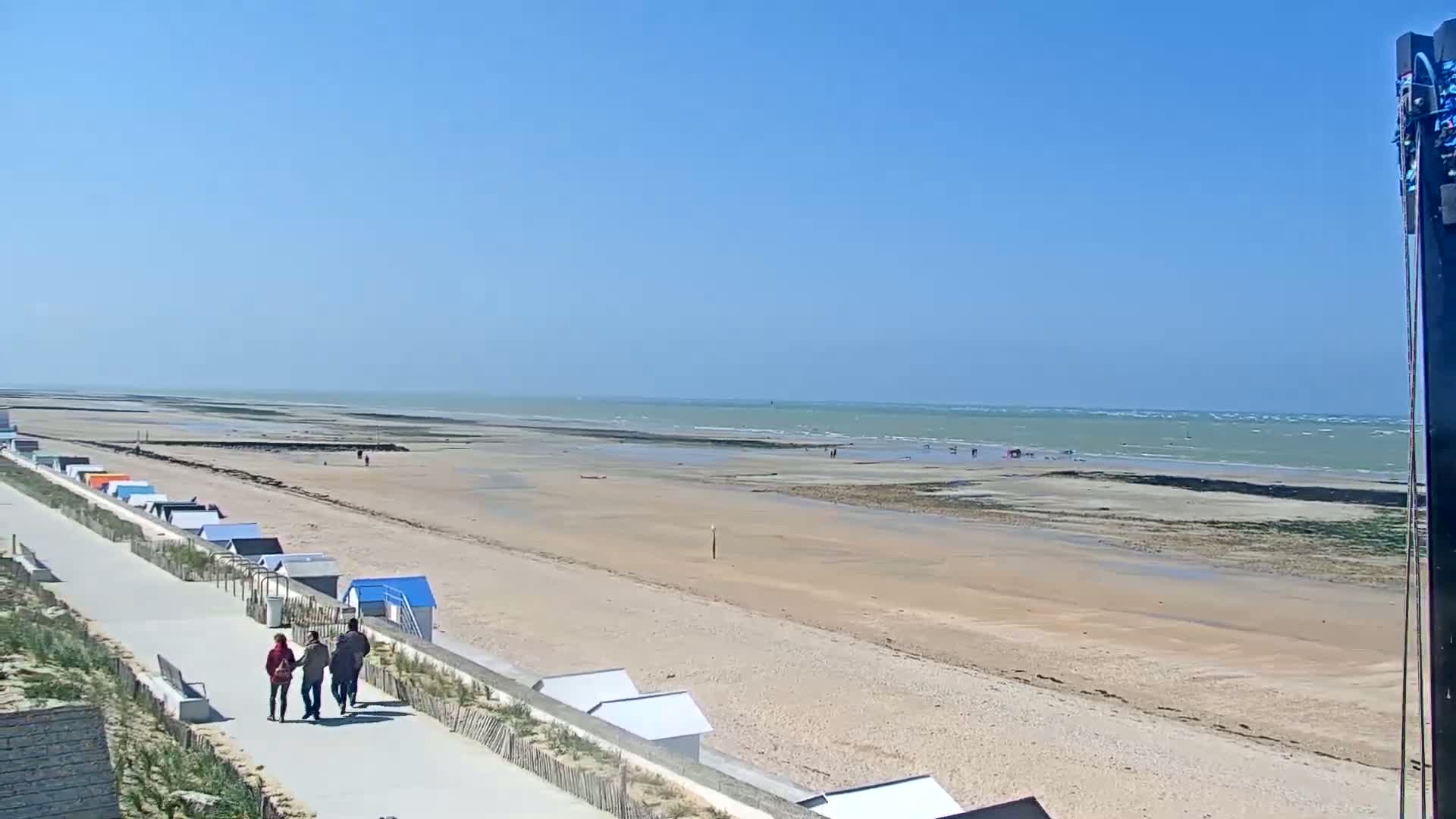 A sandy beach with small colorful beach huts stretches along a paved walkway, with a few people walking, under a clear blue sky and a calm sea with some small waves.