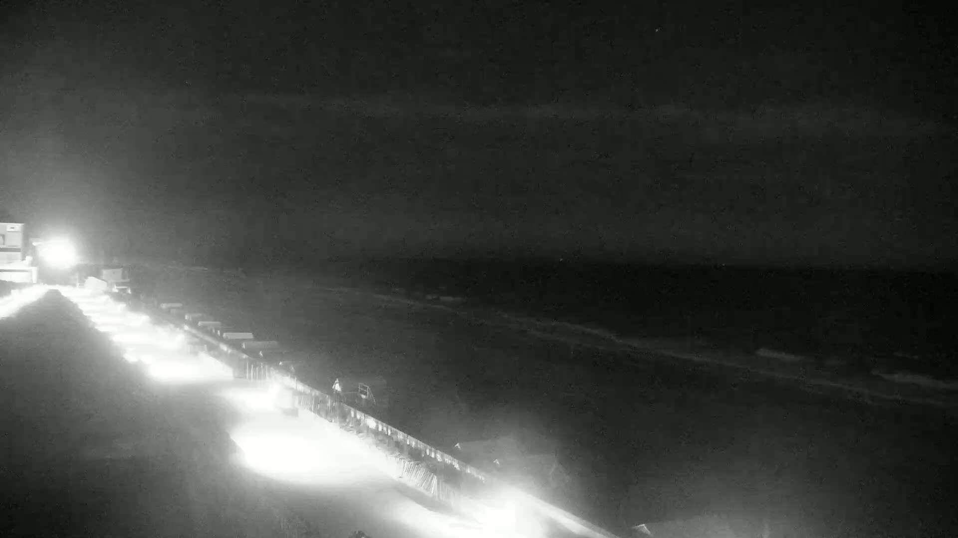 A nighttime, low-light view shows a long, linear structure with lights extending toward a dark ocean under a cloudy sky.