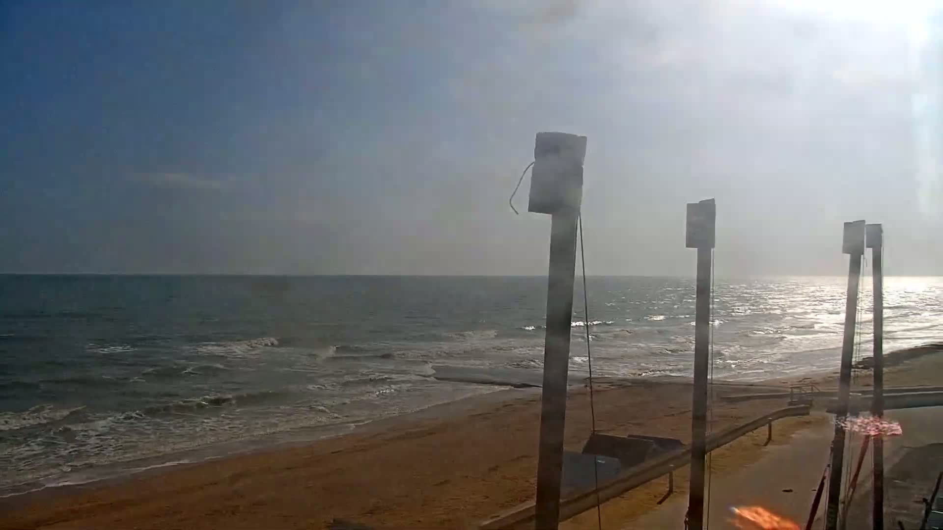 A hazy sunny day shows a sandy beach with gentle waves rolling onto the shore, near several tall posts.