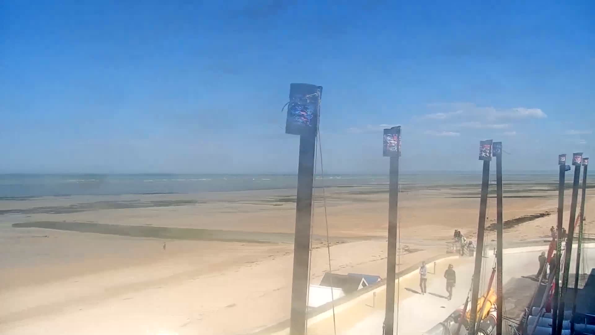 A wide sandy beach stretches to a calm ocean under a clear blue sky, with several tall poles and a few people walking along a paved area near the water.