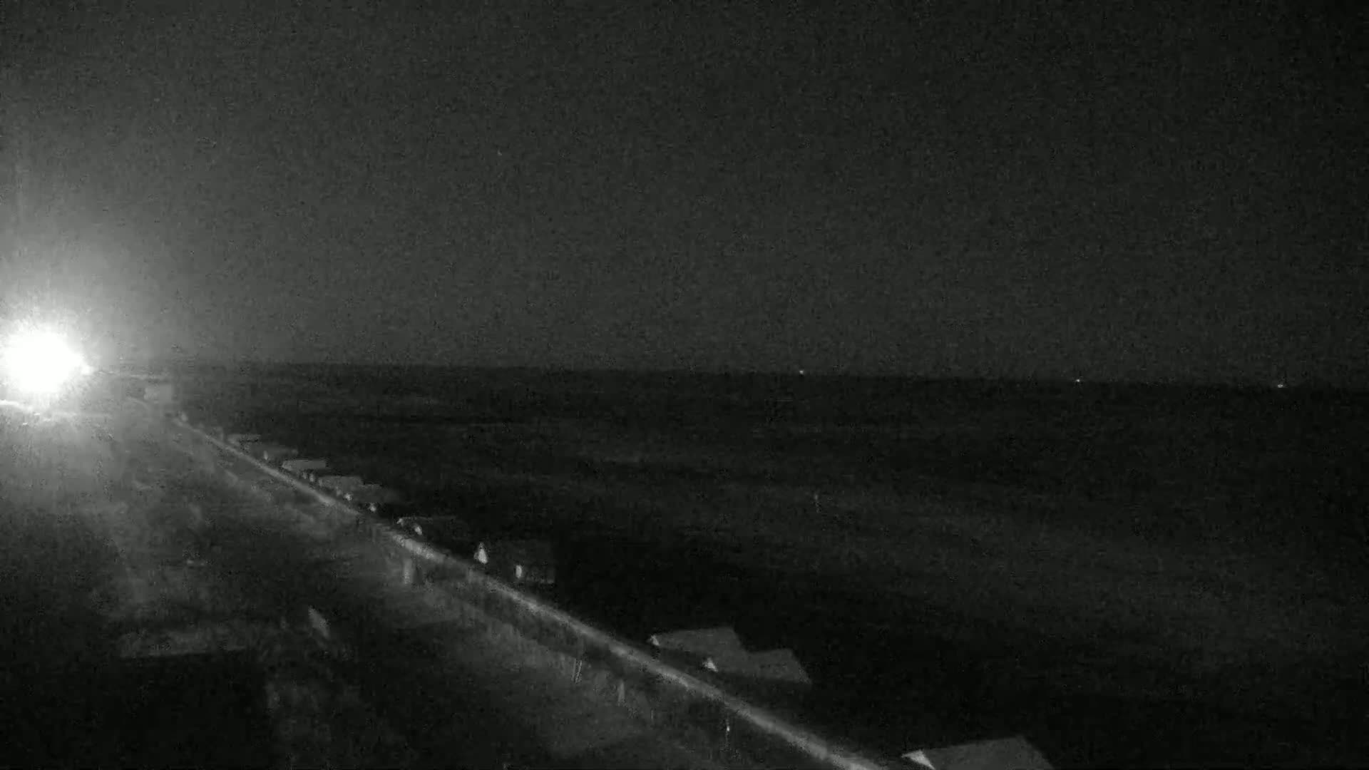 A nighttime, low-light view shows a road with vehicles driving toward the camera, alongside a structure, and a dark, flat landscape beyond.