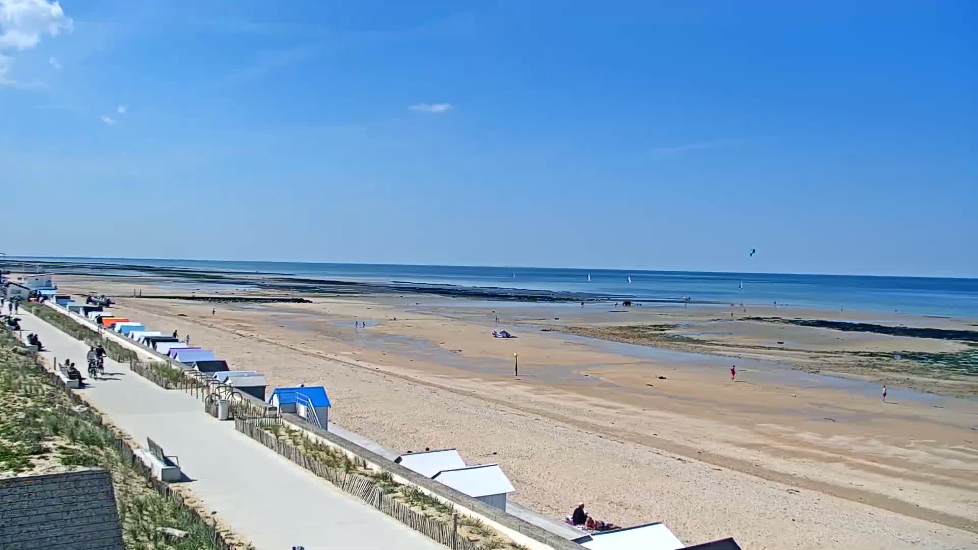 A sandy beach stretches to a calm, blue ocean under a clear, sunny sky, with colorful beach huts lining a paved walkway.